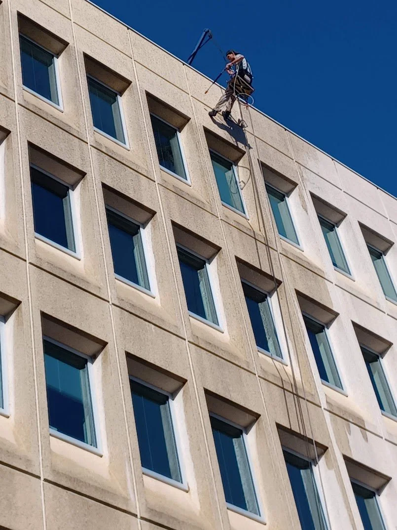A worker in safety gear cleaning windows on the upper floors of a modern building with a hydraulic lift under a clear blue sky.