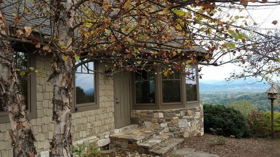 View of a house with beige siding and stone accents, surrounded by trees with autumn leaves, overlooking a distant landscape and mountains.