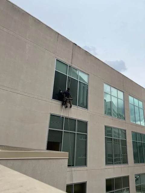 A person cleaning windows on the exterior of a modern building from a harness and rope.
