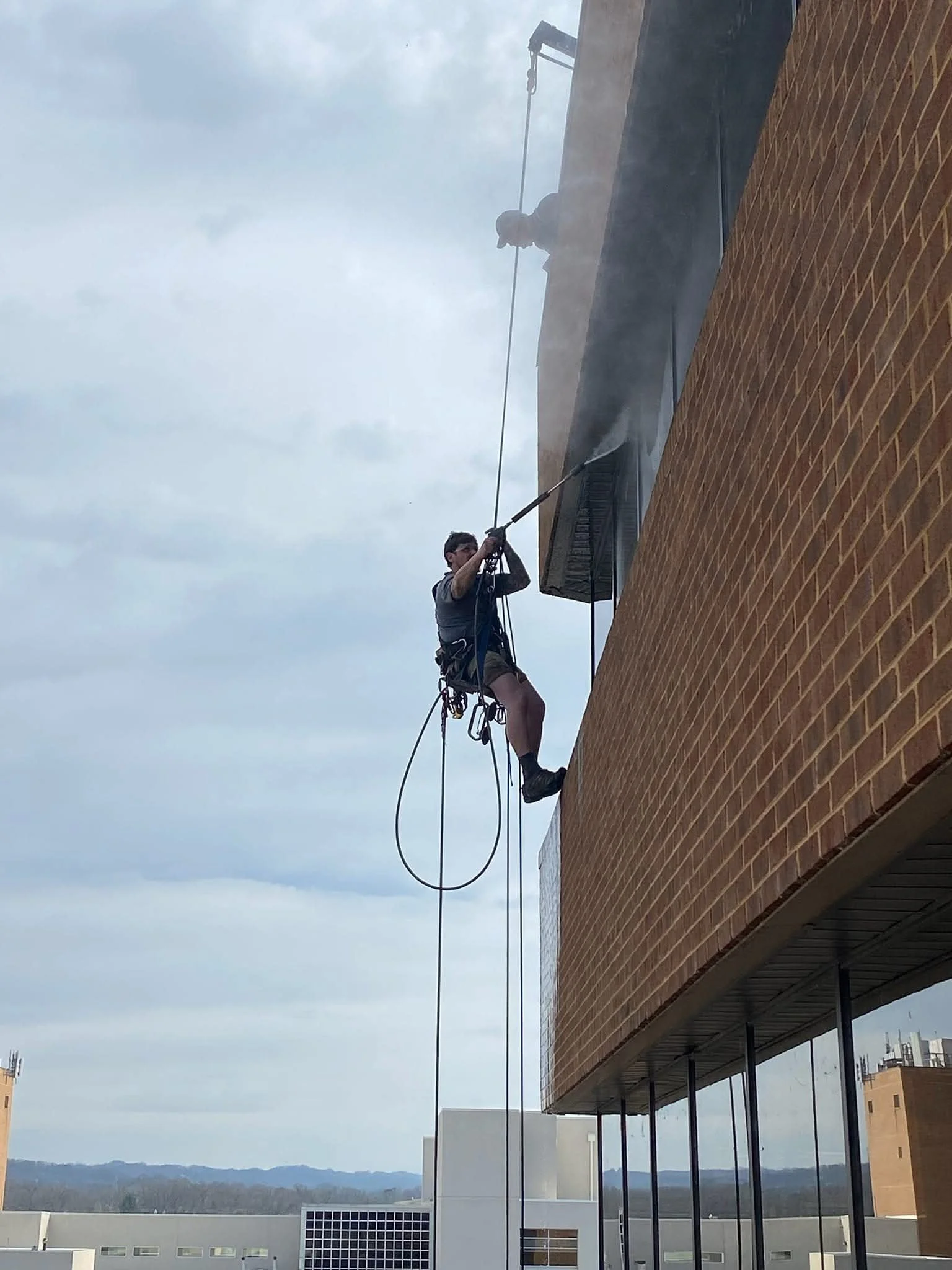 A worker cleaning the exterior window of a building with a high-pressure water hose while suspended from the building's side.