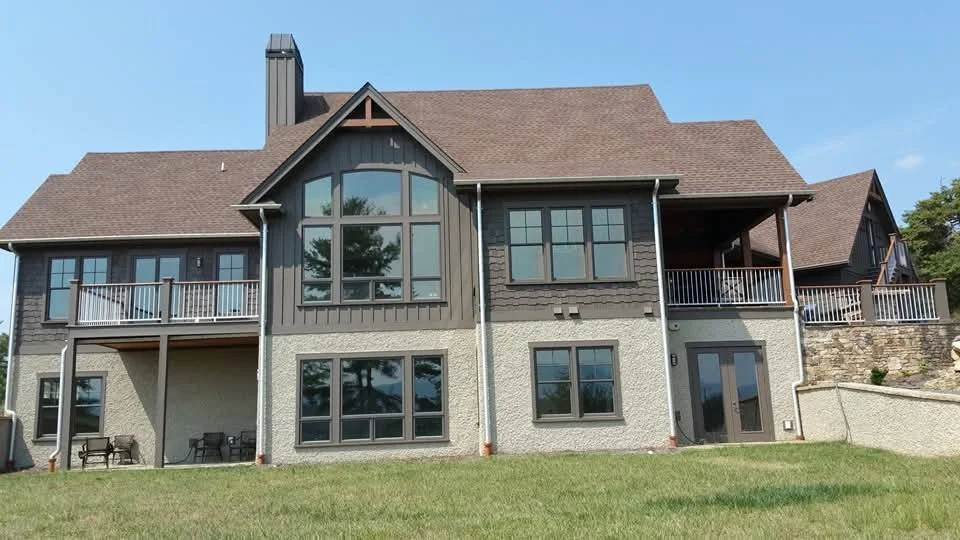 Large two-story house with a brown roof, gray and dark gray exterior, and multiple windows. It has balconies with railings on each side and a patio area below.