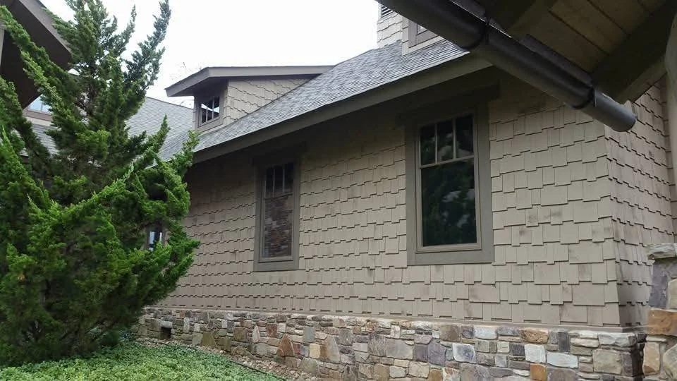 Side view of a beige house with two windows, stone foundation, and a tree next to it.