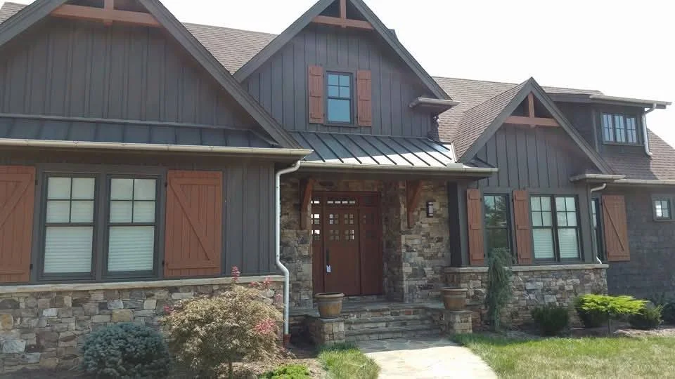 A large modern house with stone and dark siding exterior, brown shutters, and a front porch with steps leading up to the door, surrounded by a well-maintained lawn and shrubs.