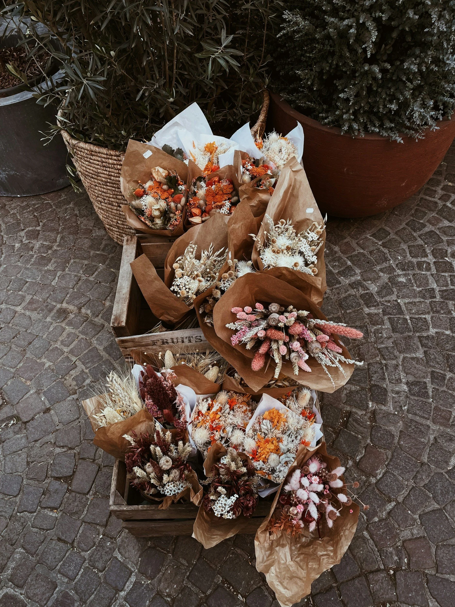 A display of various dried flower bouquets wrapped in brown paper, arranged in a wooden crate on a cobblestone surface, with large potted plants in the background.