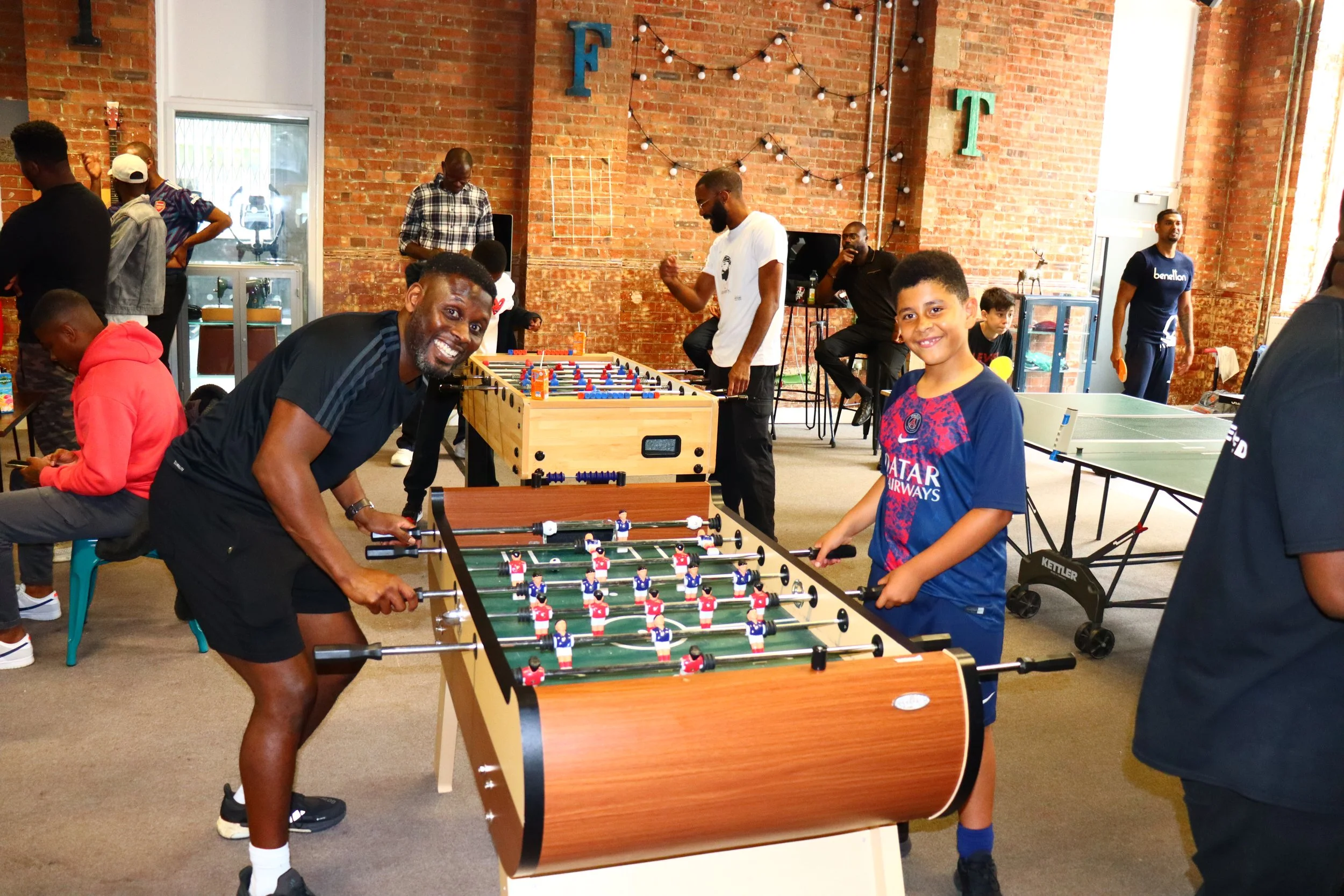 People playing foosball and enjoying themselves in an indoor recreation space with brick walls and string lights.