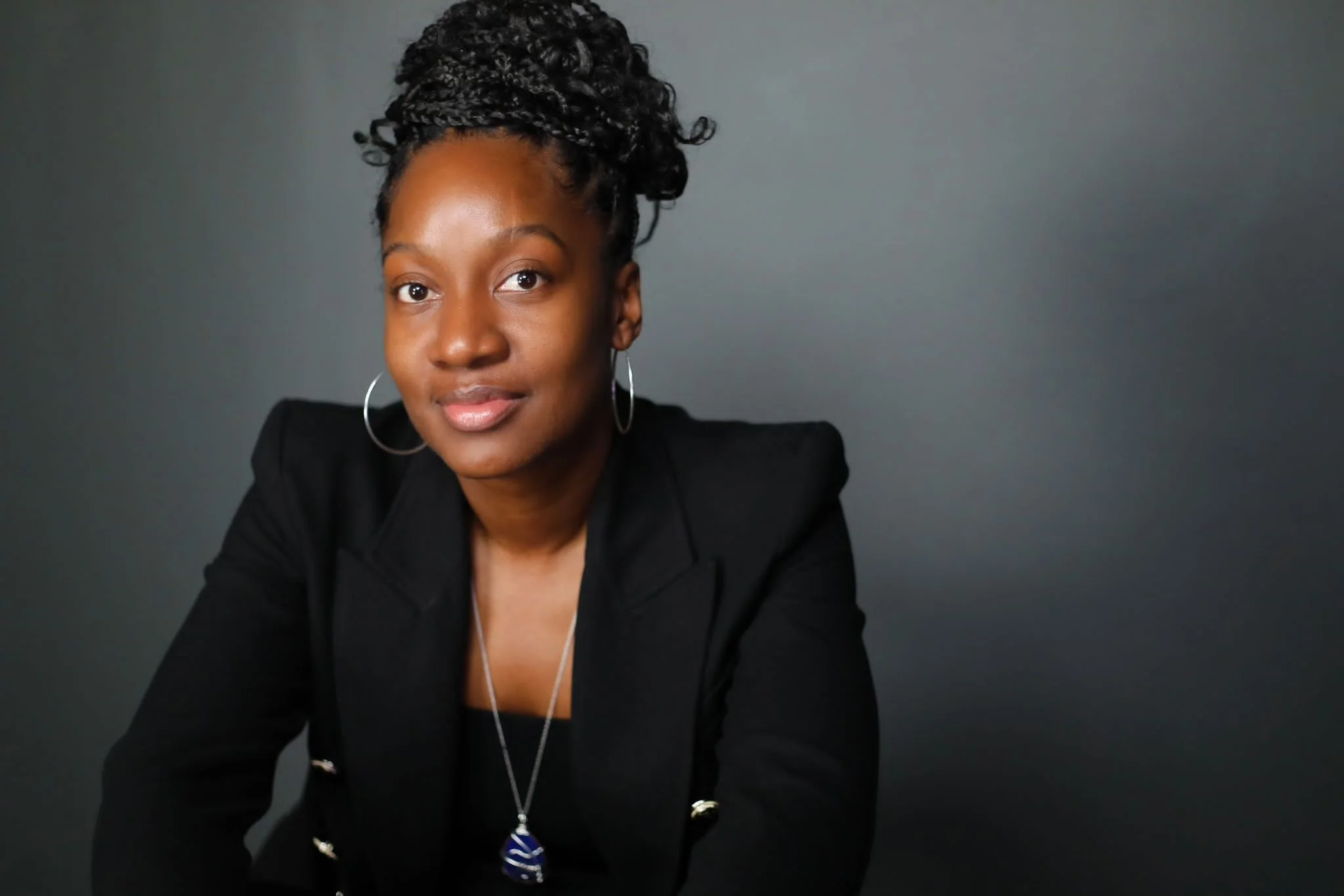 A woman with braided hair styled in an updo, wearing hoop earrings, a black blazer, and a necklace with a blue pendant, sitting against a dark gray background.