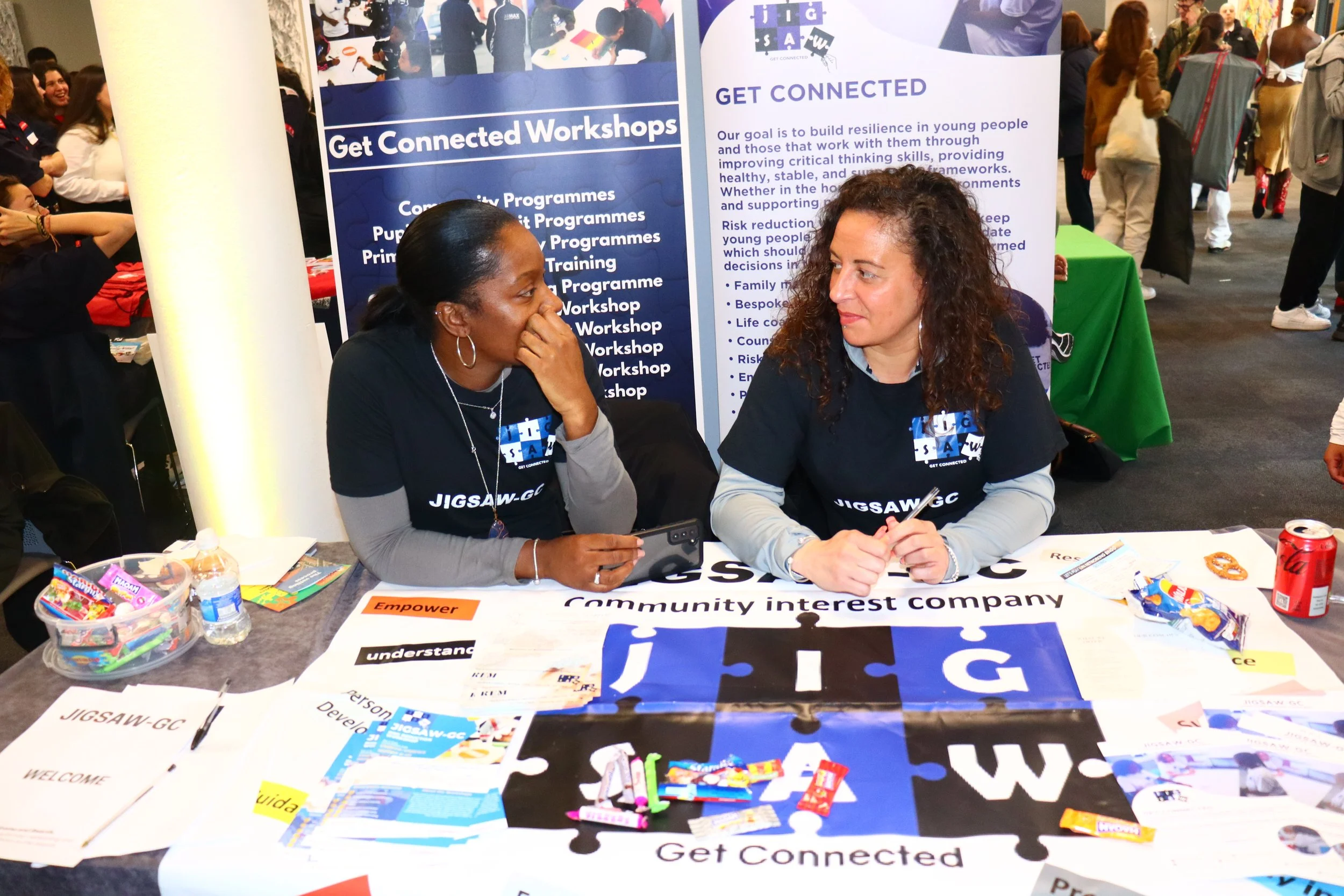 Two women sitting at a table, engaged in conversation at an event, with informational posters and promotional materials about JIGSAW-GC behind them.