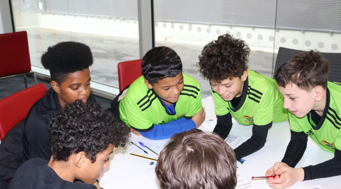 A diverse group of young boys, some with curly hair and one with afro, are gathered around a table in a modern room with glass walls, working together on a project, smiling and engaged.