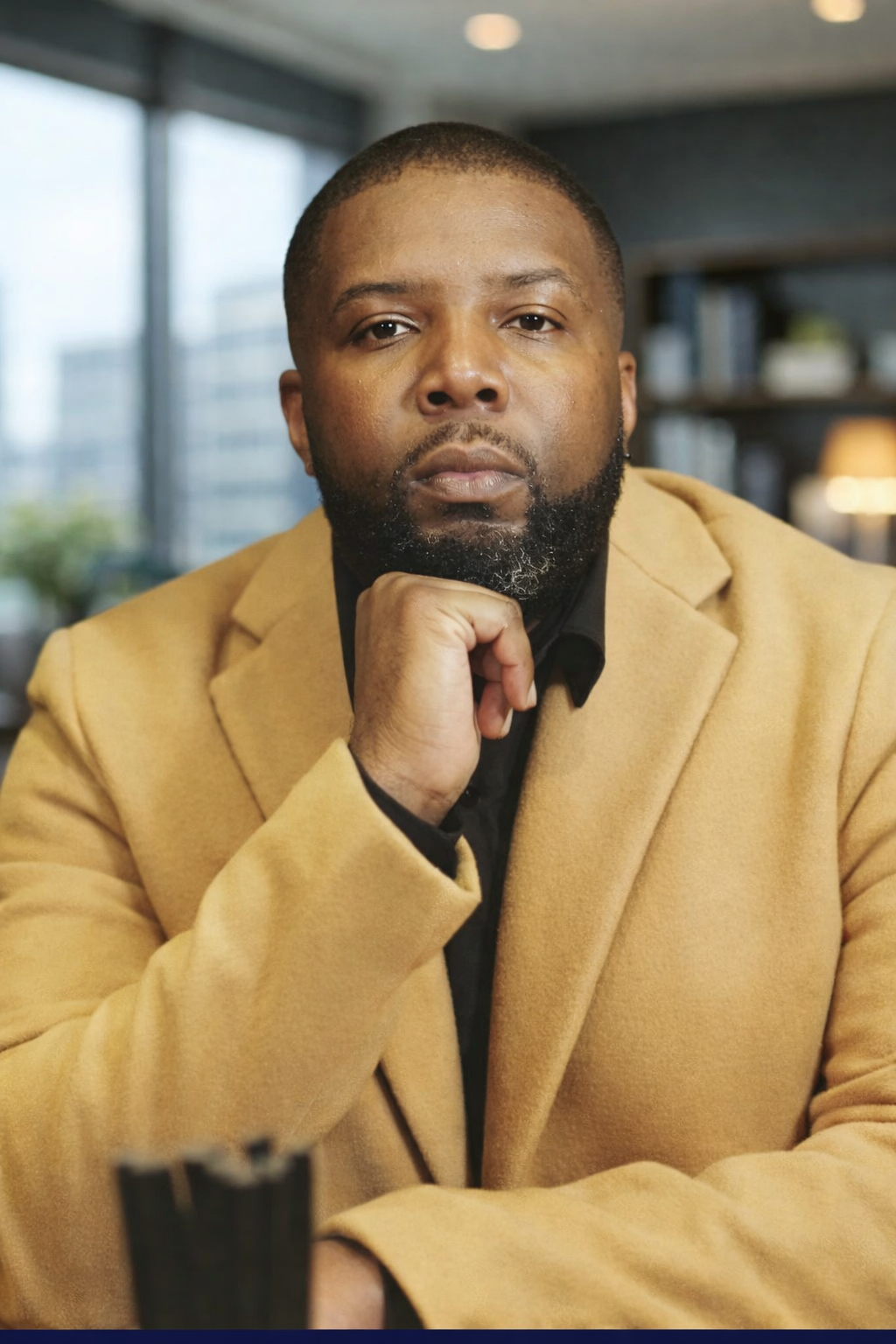 A man with a beard wearing a tan blazer and black shirt sitting indoors, with a thoughtful expression and resting his chin on his hand.