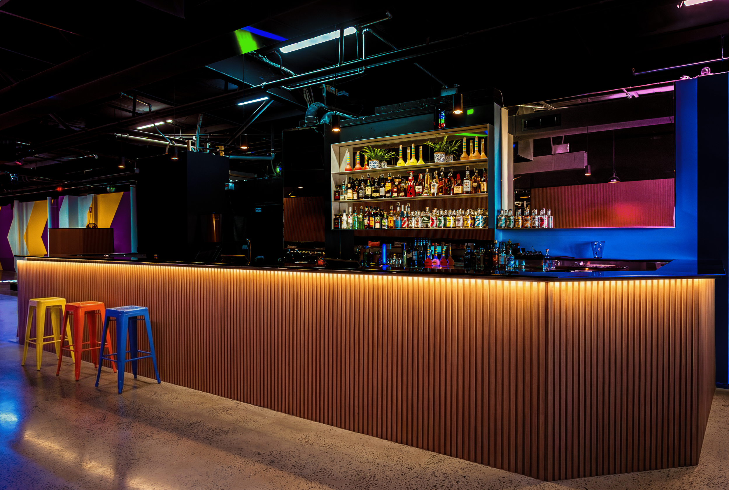 A modern bar with a wooden front counter, colorful stools, and a well-lit shelf of liquor bottles behind the bar, illuminated by decorative lighting and neon accents.