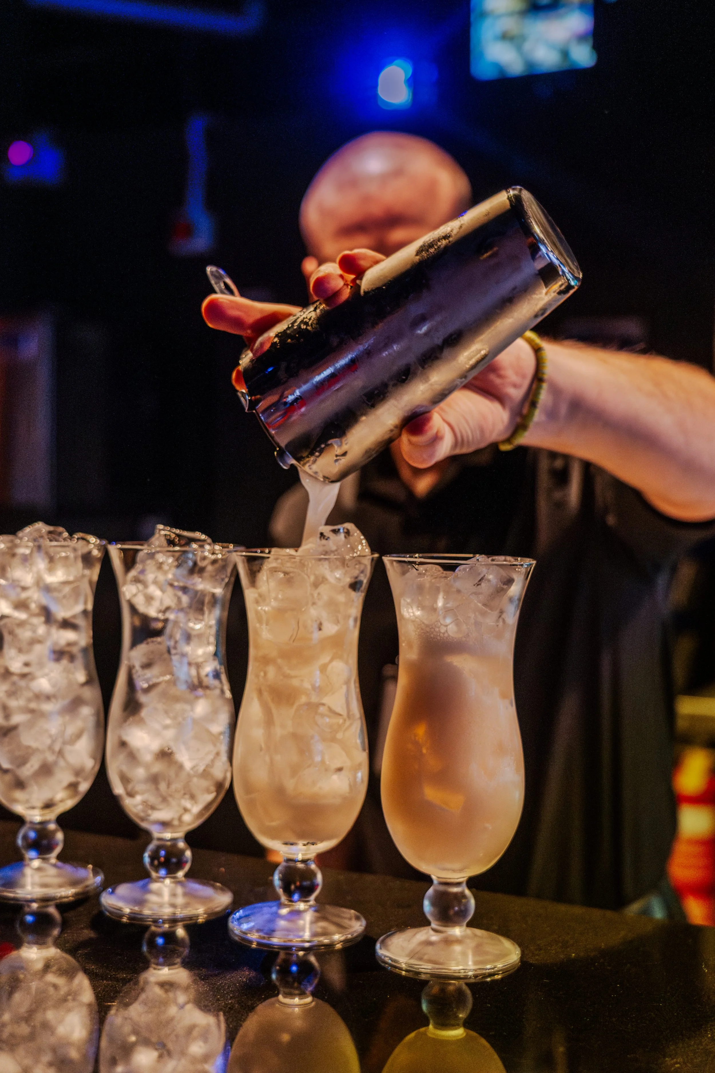 Bartender pouring a creamy cocktail into tall glasses filled with ice at a bar.