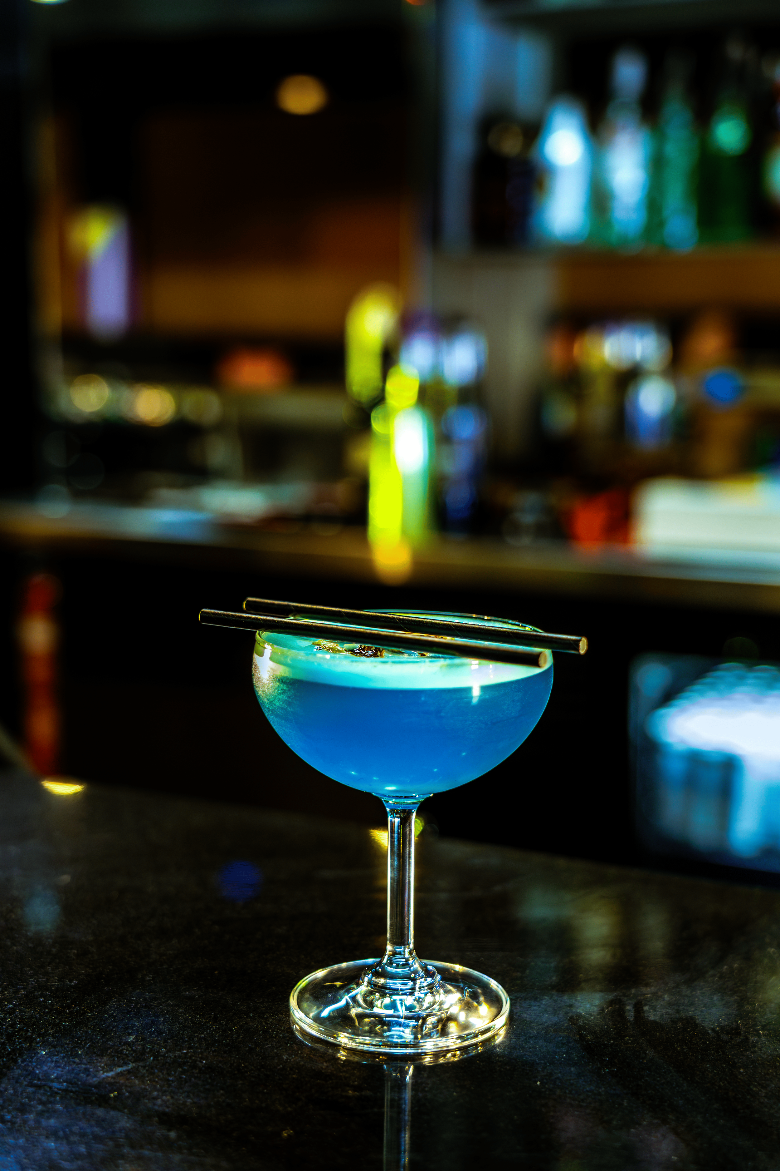 A shot of a blue cocktail in a stemmed glass garnished with metallic chopsticks on top, on a bar counter with a blurry bar background.