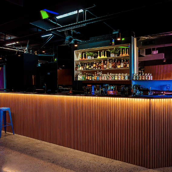 Modern bar with illuminated wooden counter, backlit shelves with various bottles and glasses, and contemporary lighting fixtures in a dimly lit setting.