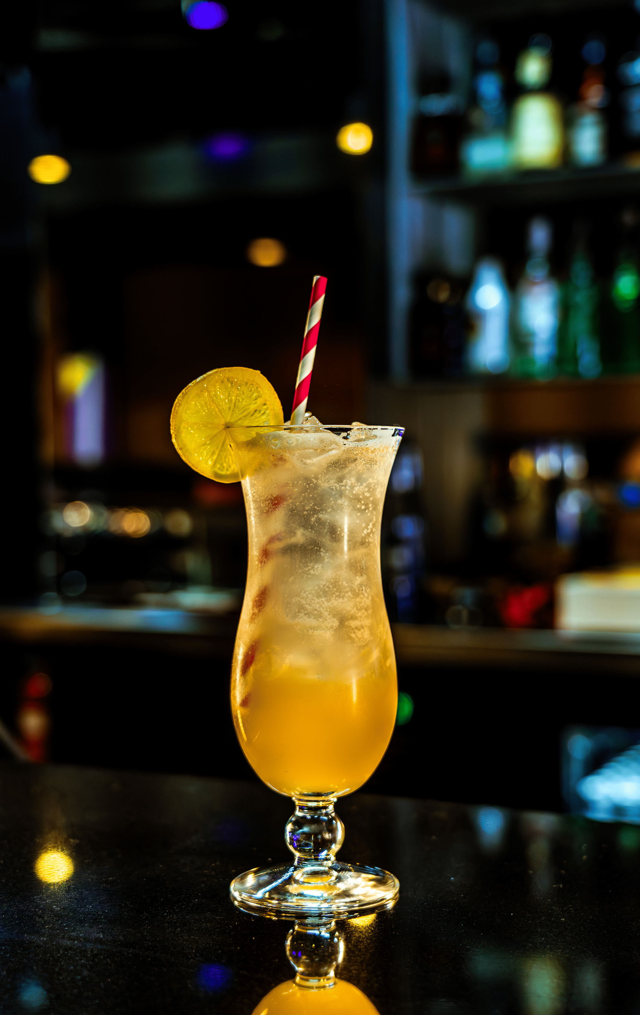 A tall, curved glass filled with a yellow cocktail garnished with a lemon slice and a red-and-white striped straw, placed on a dark reflective surface with a blurred bar background.