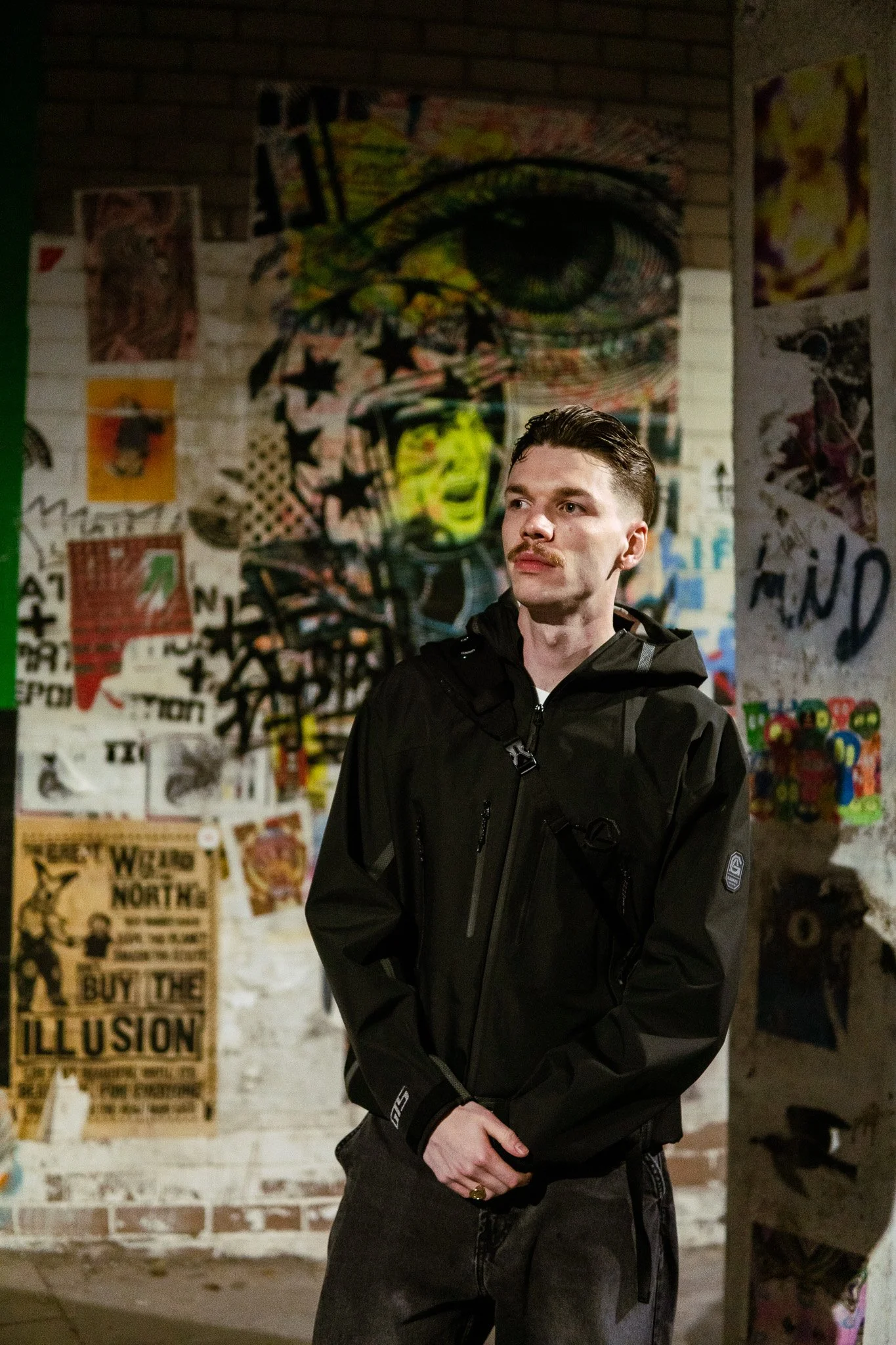 A young man with a mustache and slicked-back hair standing in front of graffiti art on a brick wall, wearing a black jacket and dark pants.