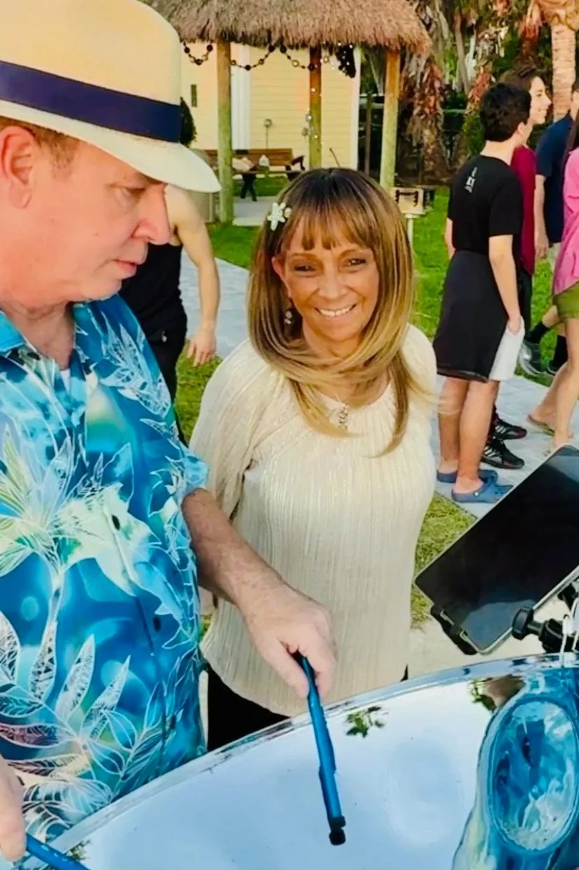 A man in a blue Hawaiian shirt and a woman in a beige top standing outdoors at a gathering. The man is playing a steel drum for a wedding at Sunset Beach (also known as Sunset Park Beach) in Key Colony Beach, Florida. In the Florida Keys.