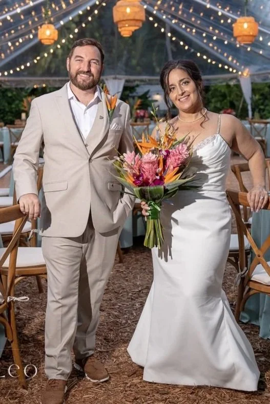 A bride and groom standing inside a decorated wedding reception area with string lights and orange paper lanterns, the bride holding a large bouquet of pink and orange flowers, both smiling at the camera. Hemingway House and Museum, Key West, Florida