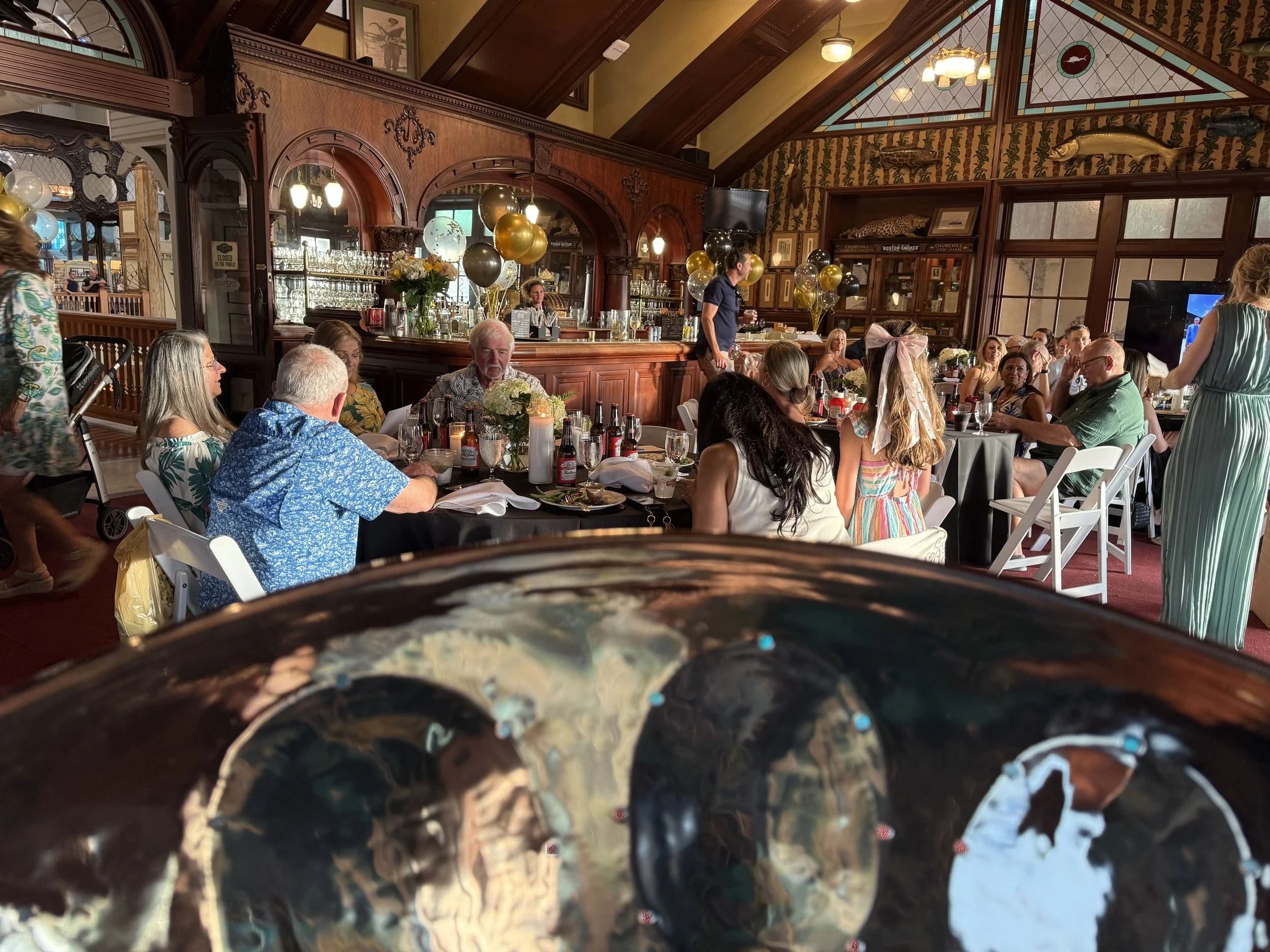 A gathering at a restaurant with people seated at tables decorated with balloons and flowers, and a bar area in the background. Islamorada Fish Company Restaurant, Live steel drum Music for a birthday party. 