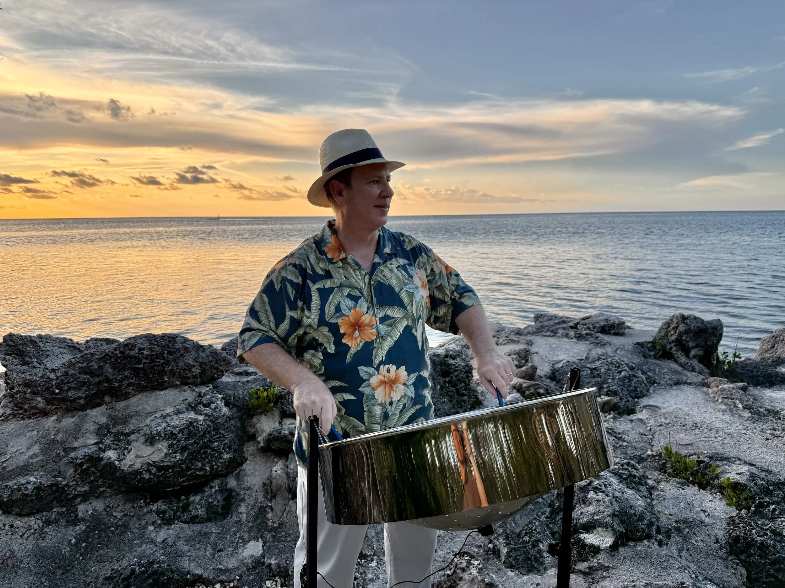 A man in a Hawaiian shirt and Panama hat stands by the rocky shoreline playing a steel drum, with a sunset over the ocean in the background. Crane Point Hammock, Marathon Florida, Florida Keys.
