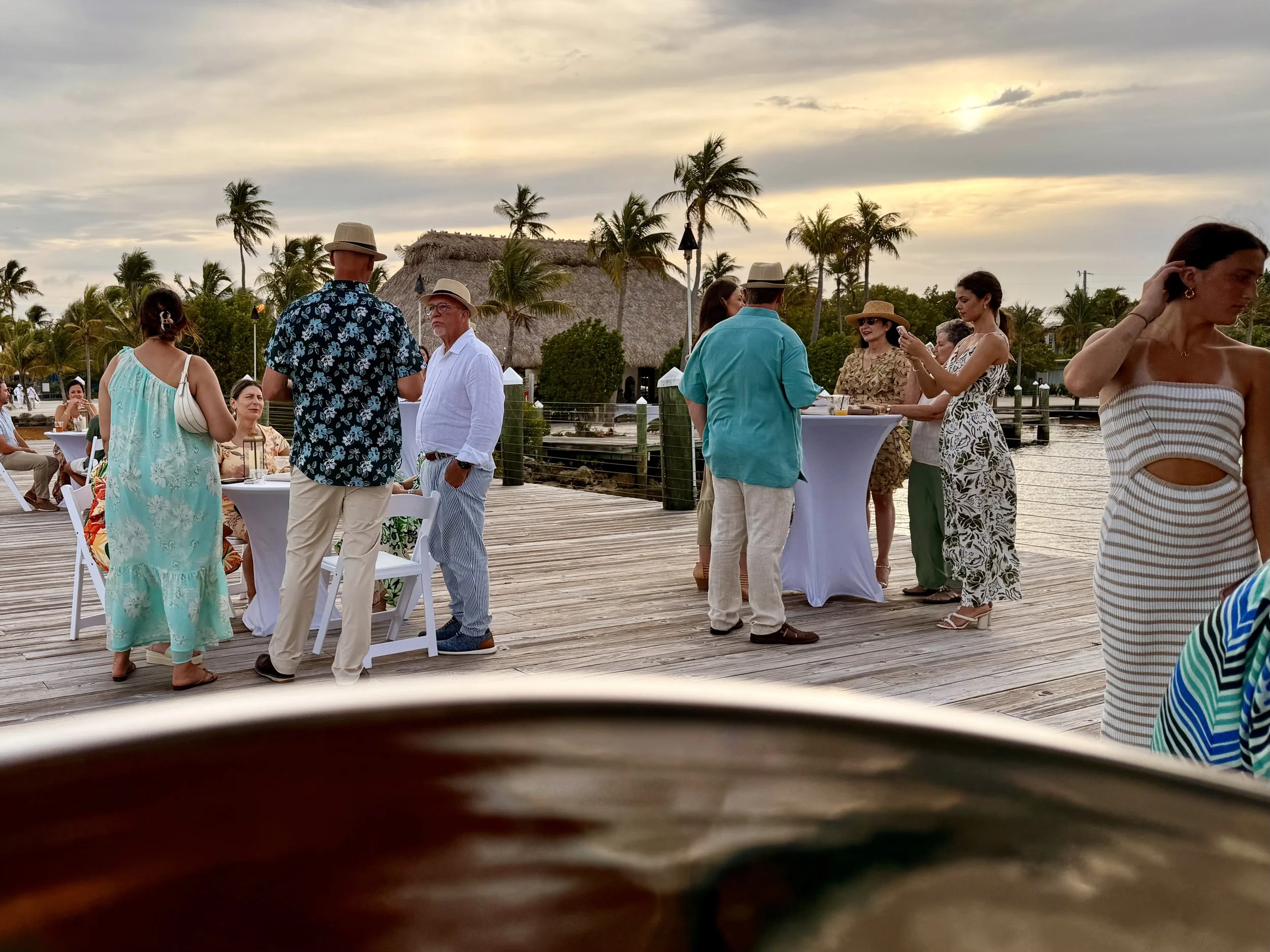 People at an outdoor gathering for a wedding cocktail hour on a wooden deck with palm trees and a thatched-roof structure in the background at sunset, socializing and taking photos. Three Waters Resort, Islamorada Florida, Florida Keys