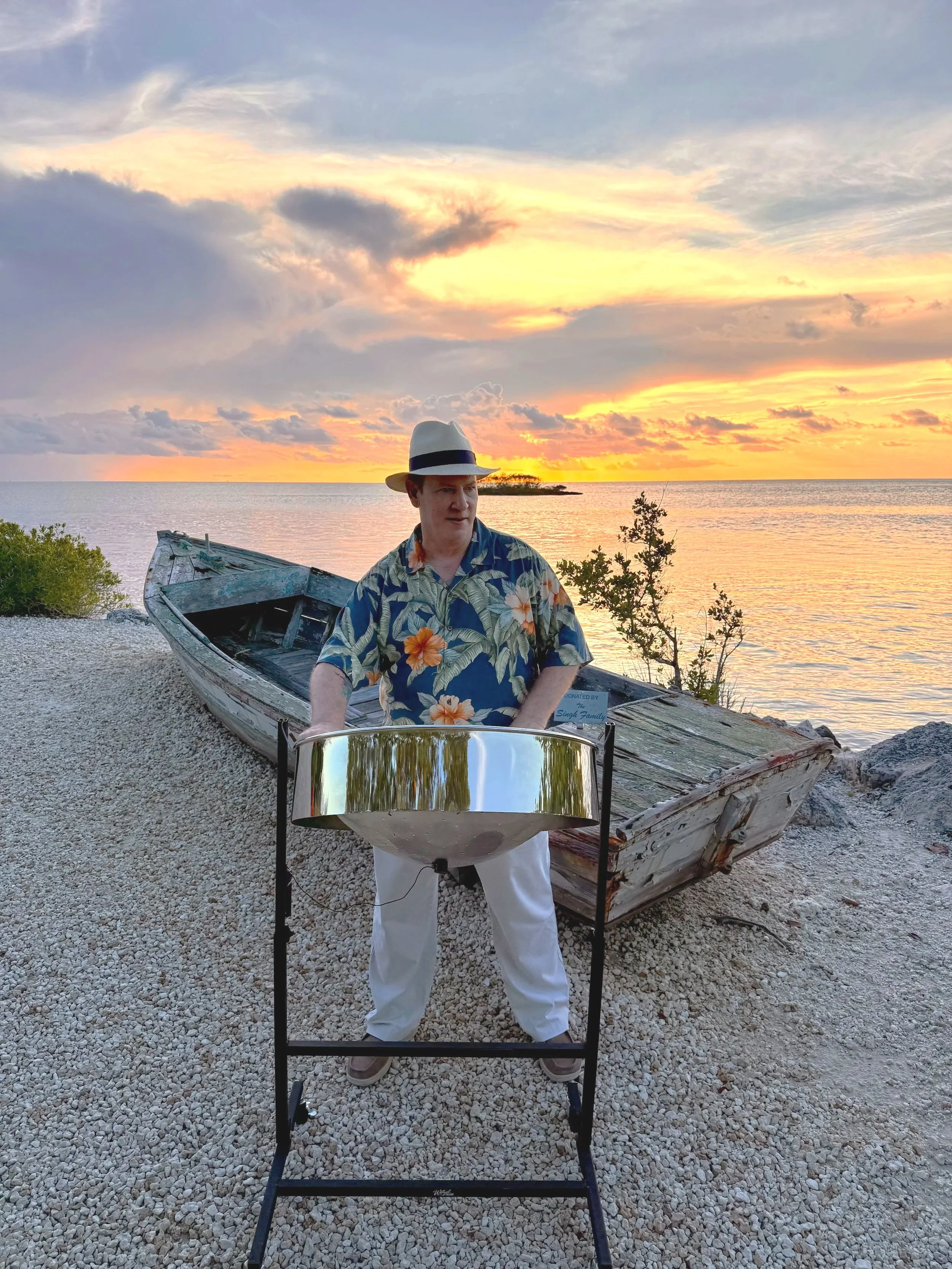A man wearing a tropical shirt and a straw hat playing live steel drum music for a private event outdoors near water and green bushes. Crane Point Hammock, Marathon Florida, Florida Keys.