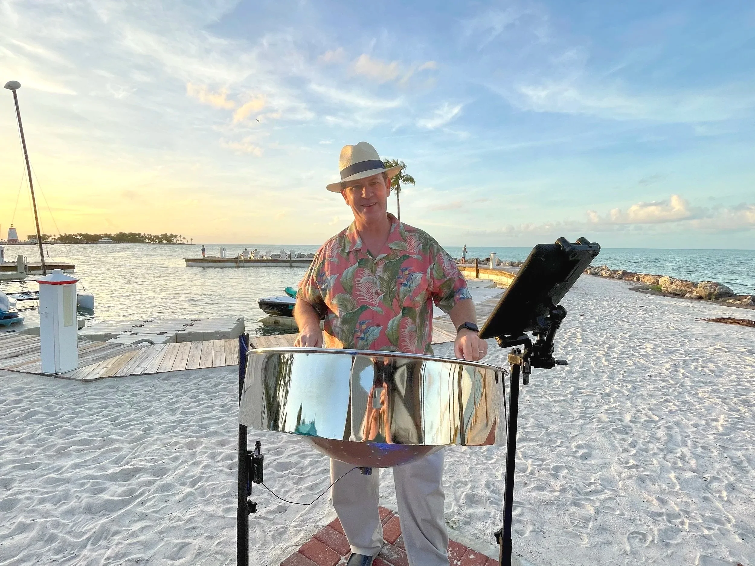 A man wearing a floral shirt and a fedora hat playing live steel drum music on a sandy beach during sunset, with a marina and calm ocean in the background. Tranquility Bay Beach Resort, Marathon Florida, Florida Keys.