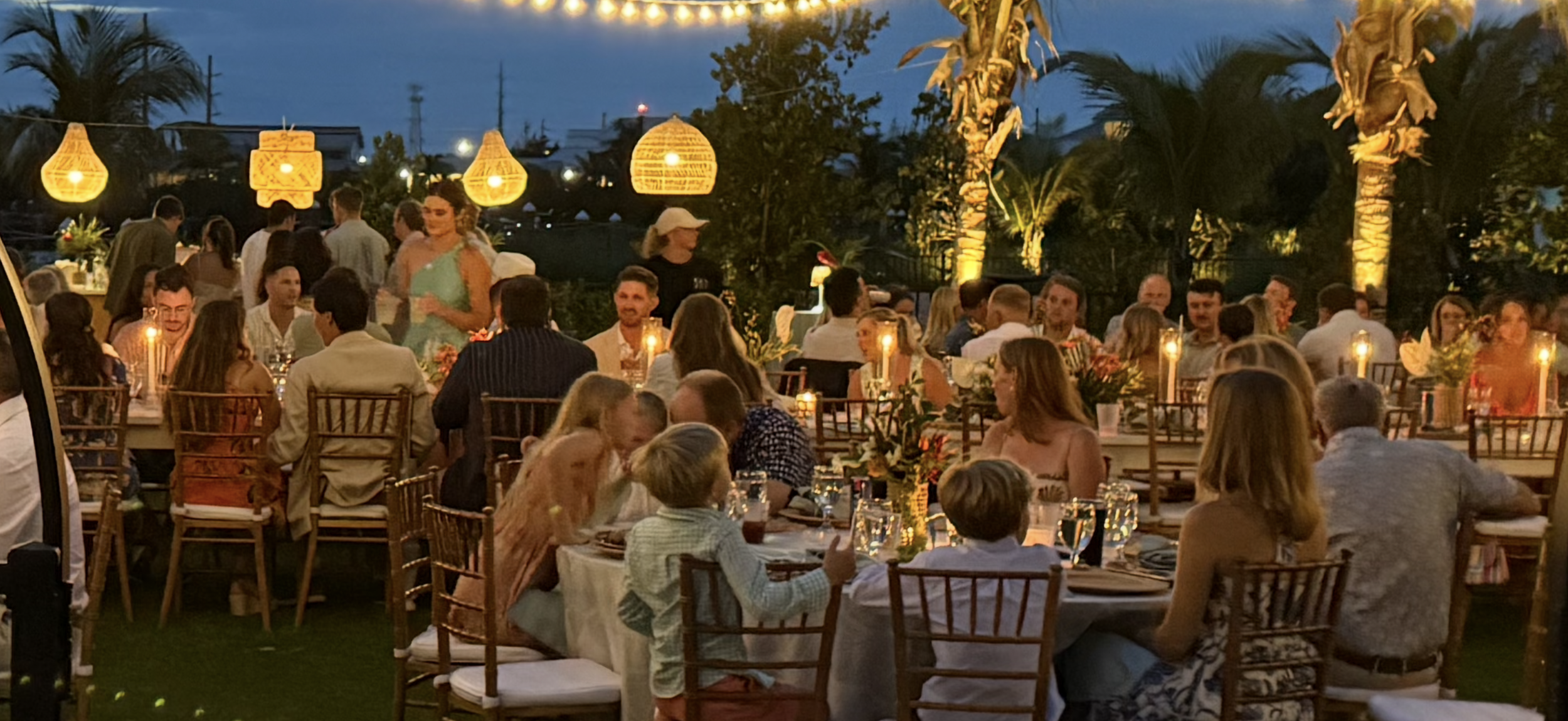 An outdoor evening wedding reception dinner event with live steel drum music, string lights, palm trees, and many guests seated and standing around tables with candles, in a tropical setting. Lunara Bay, Key West Florida, Florida Keys,