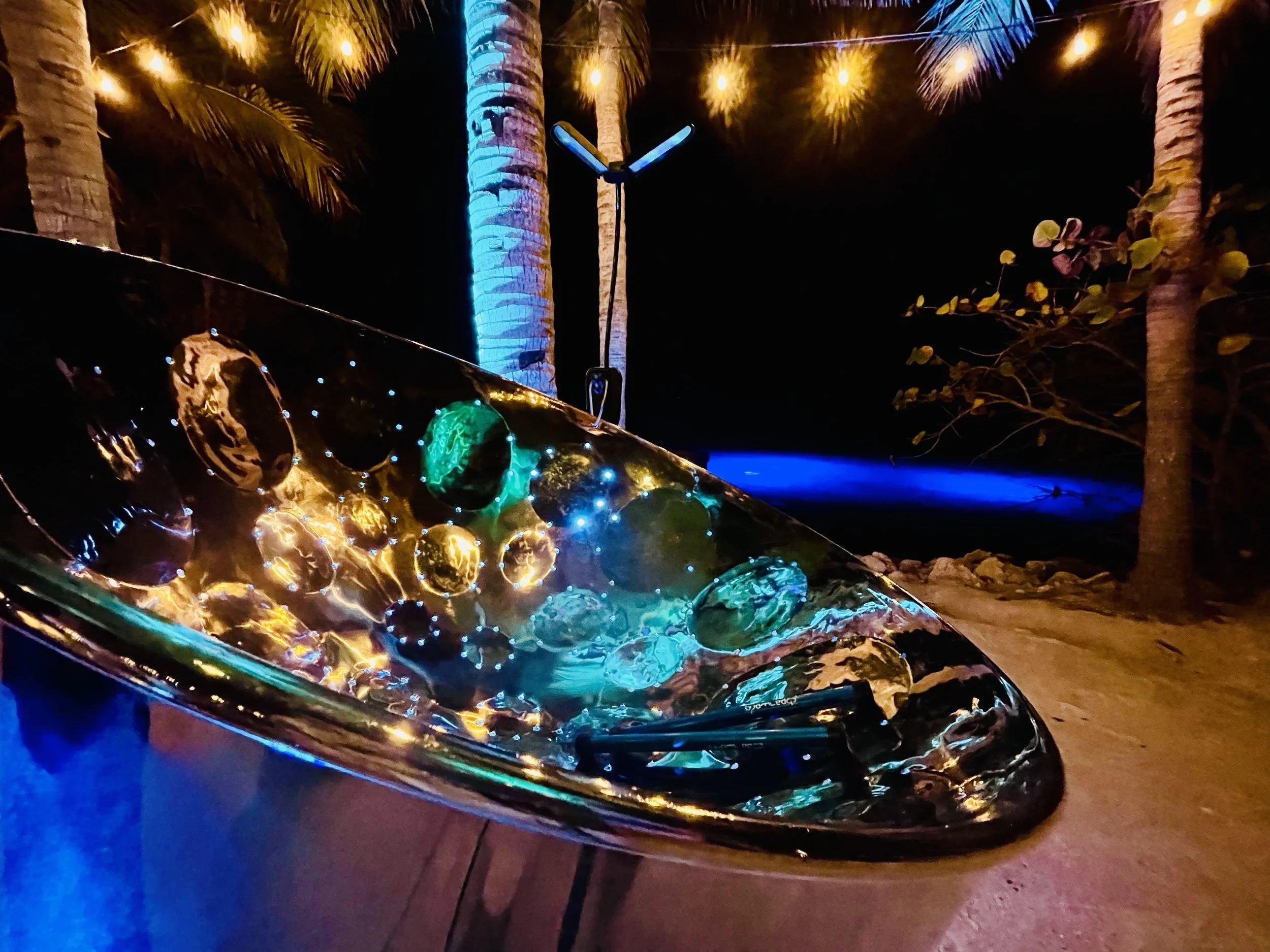 A shimmering steel drum, lit at night. Lights reflecting on the surface, surrounded by tall palm trees illuminated with blue lights at night. Audubon House and Tropical Gardens, Key West, Florida Keys