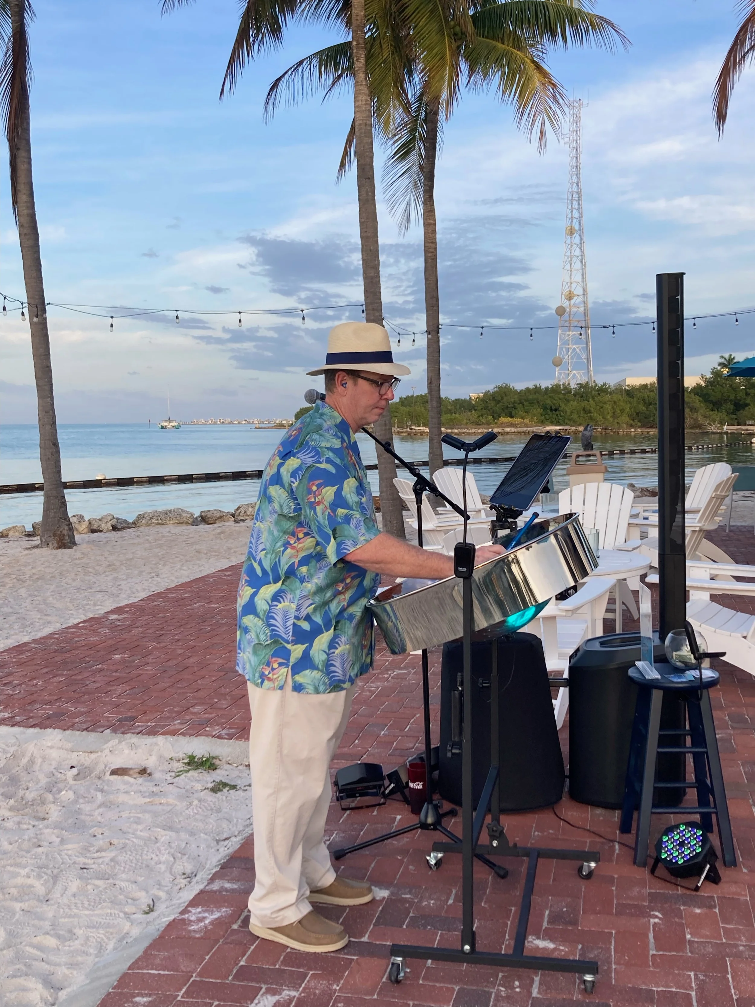 A man wearing a summer Hawaiian shirt, beige pants, a hat, and glasses is playing a steel drum by the beach during sunset. There are palm trees and a view of the ocean in the background. Faro Blanco Resort, Marathon Florida, Florida Keys.