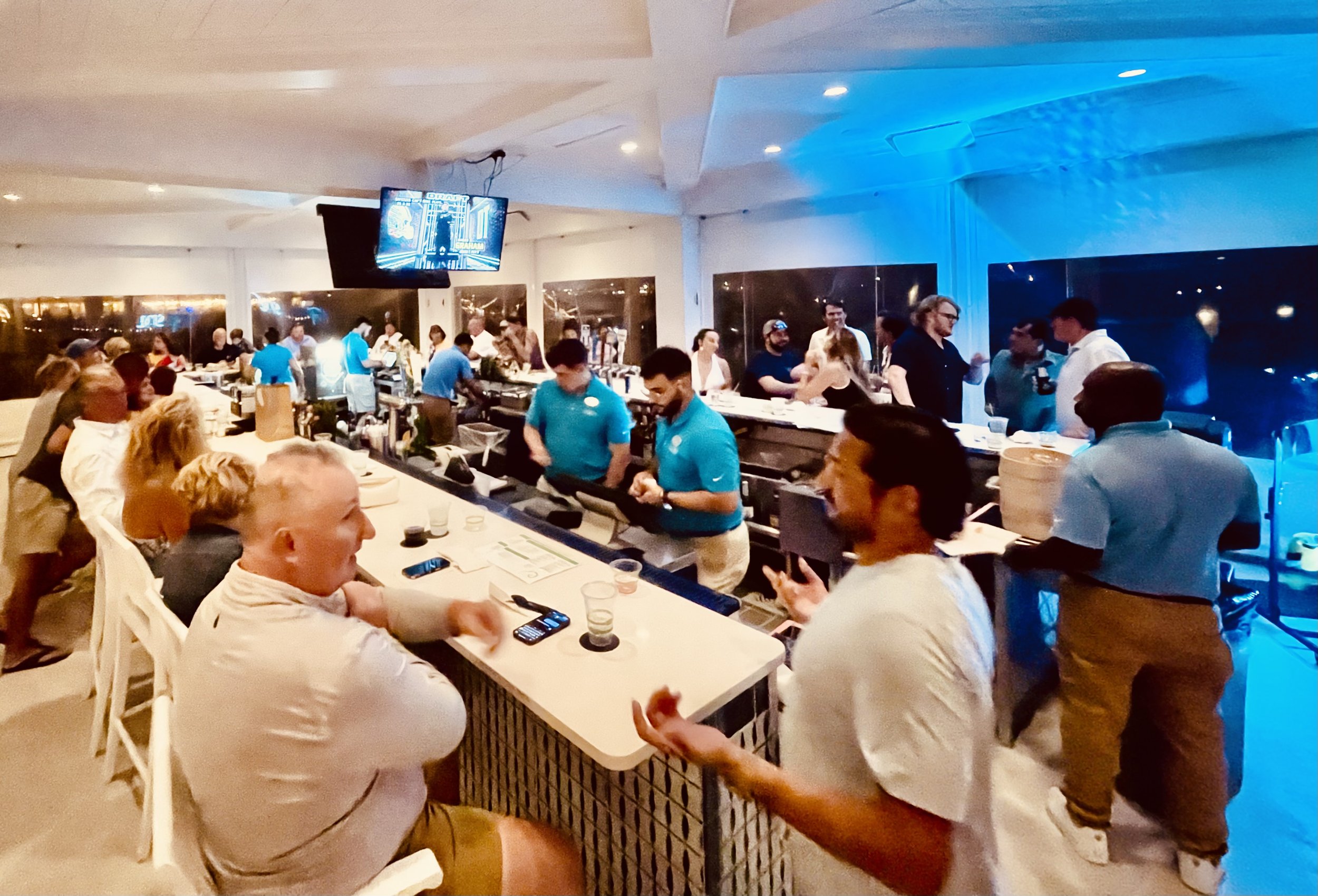 A lively bar or restaurant with people socializing, listening to live steel drum music including some seated at a counter and others standing, with a bartender preparing drinks.  Isla Bella Beach Resort, Marathon Florida, Florida Keys