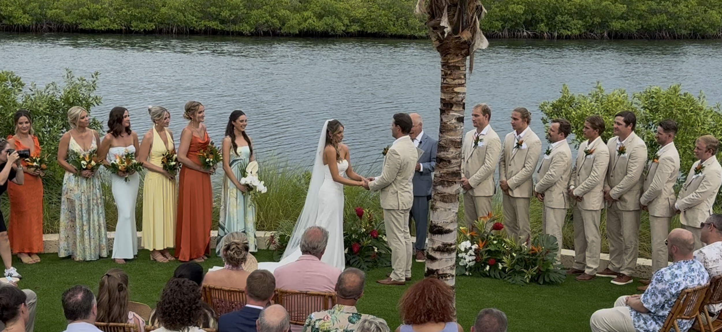 Wedding ceremony outdoors by a body of water with the bride and groom exchanging vows, surrounded by bridesmaids and groomsmen, with guests seated in front. Lunara Bay, Key West Florida, Florida Keys. Wedding Ceremony Music