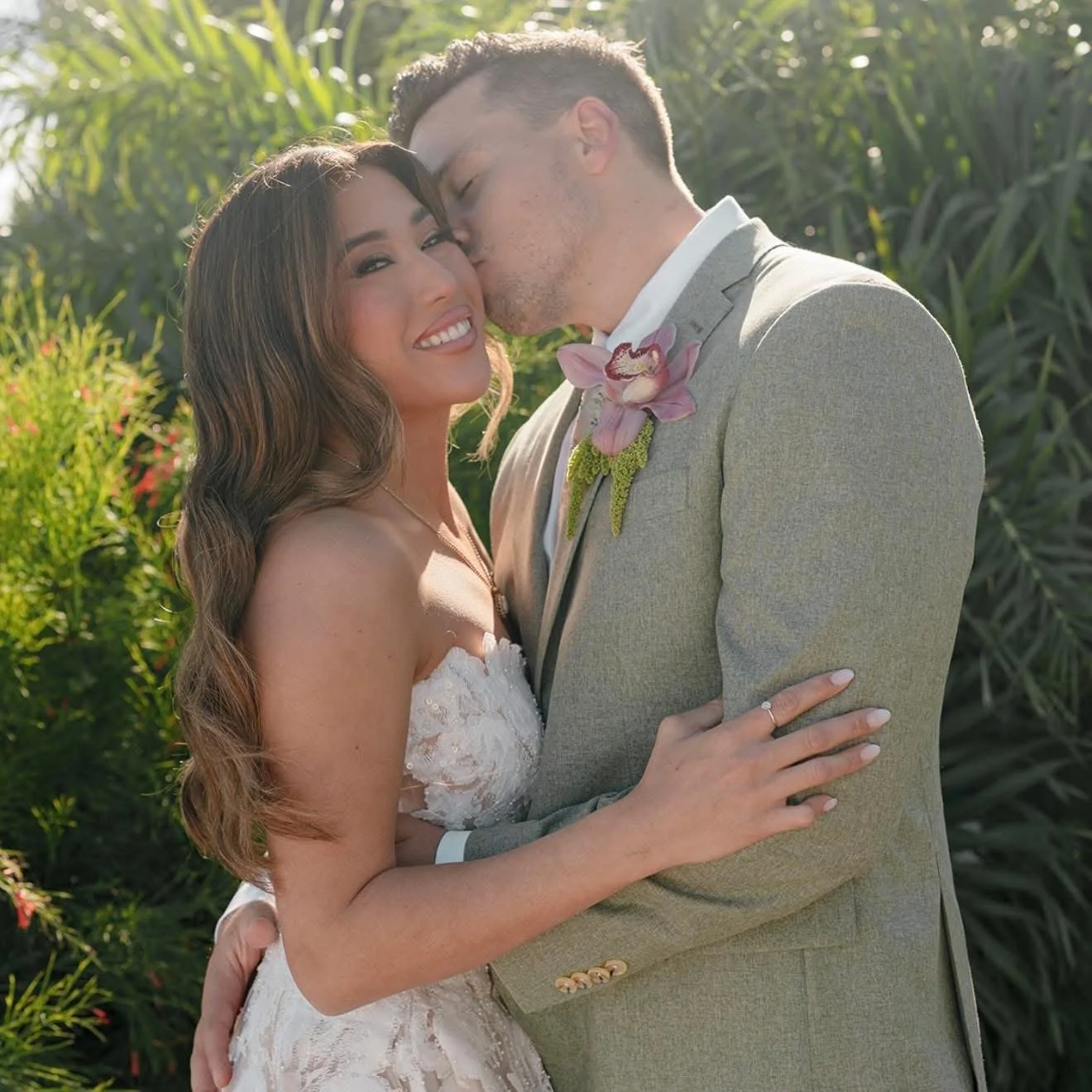 A newlywed couple sharing a kiss outdoors. The bride is smiling, wearing a white lace wedding dress, and the groom is in a light gray suit with a pink orchid boutonniere. Welcome Reception, Marker 88 Restaurant, Islamorada Florida, Florida Keys