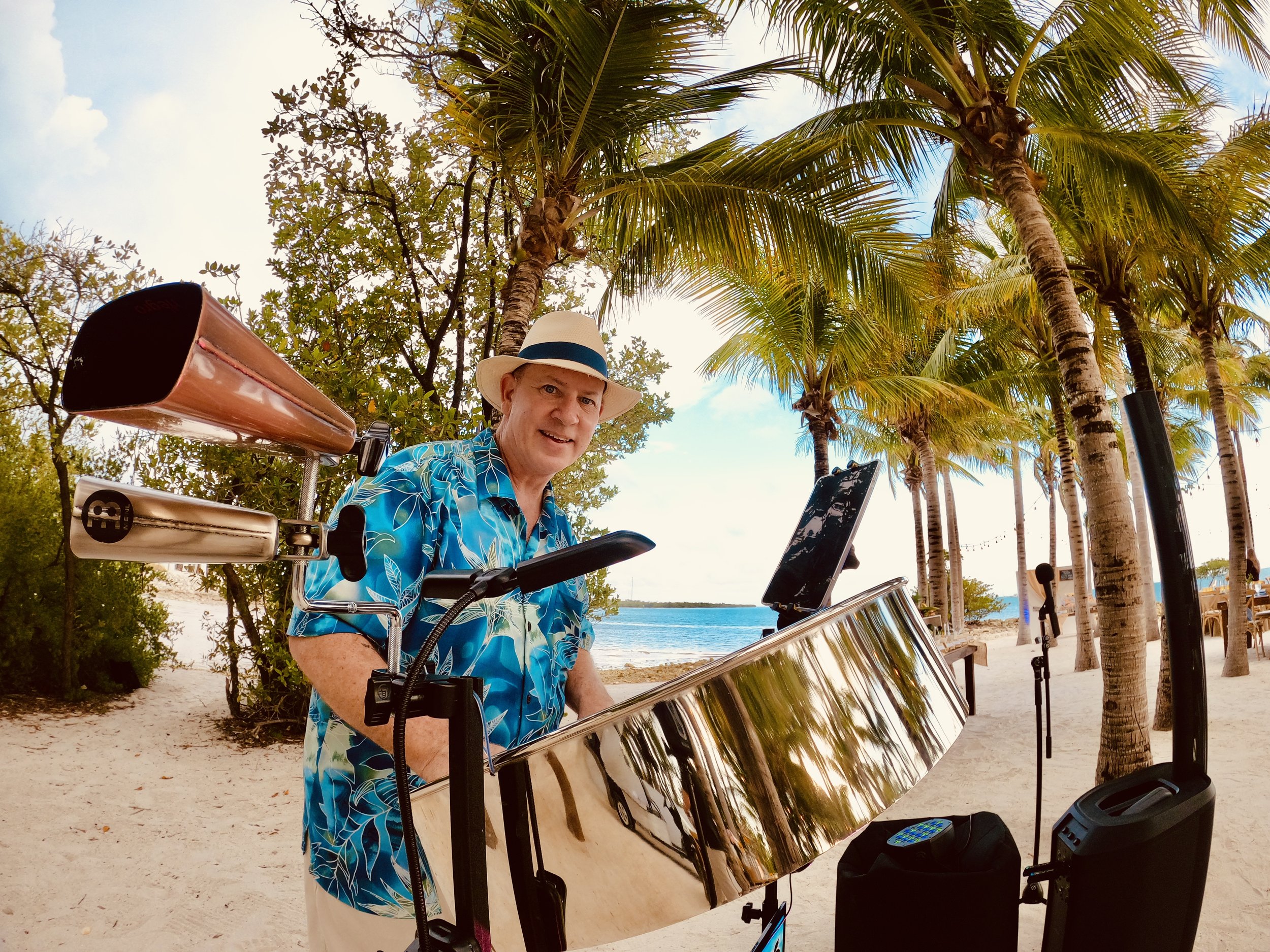 A man in a tropical shirt and hat playing a steel drum on a beach with palm trees and the ocean in the background. Live Music Event, Weekly, Isla Bella Beach Resort, Marathon Florida, Florida Keys