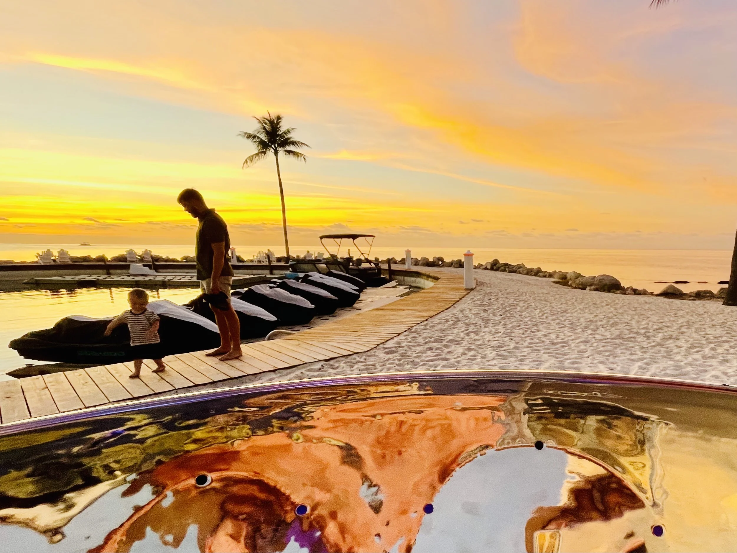 A sunset scene on a beach with a man and a child walking along a dock next to boats, with a paddleboard in the foreground and a palm tree against a colorful sky. Marathon Florida, Key West Florida, Corporate Event/Weddings, Steel drum