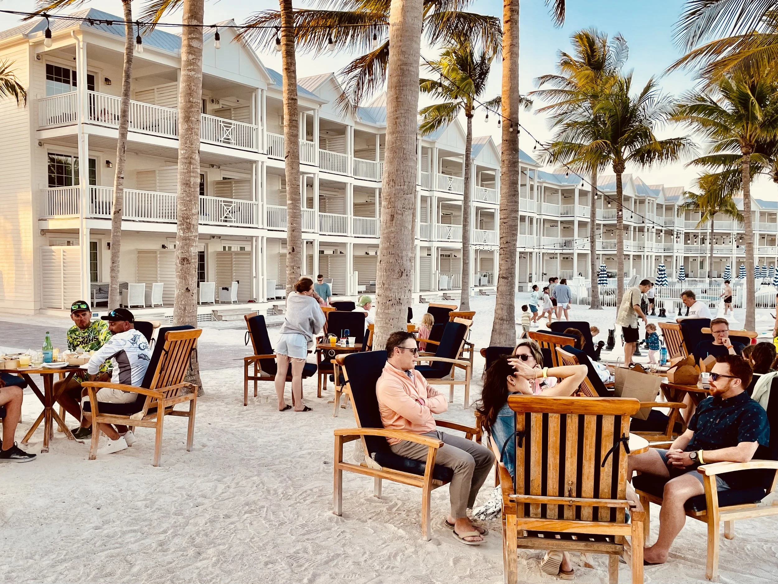 People sitting and socializing at wooden tables listening to steel drum live music with chairs on a sandy beach with high-rise buildings and palm trees in the background, during sunset. Isla Bella Beach Resort, Marathon Florida, Florida Keys
