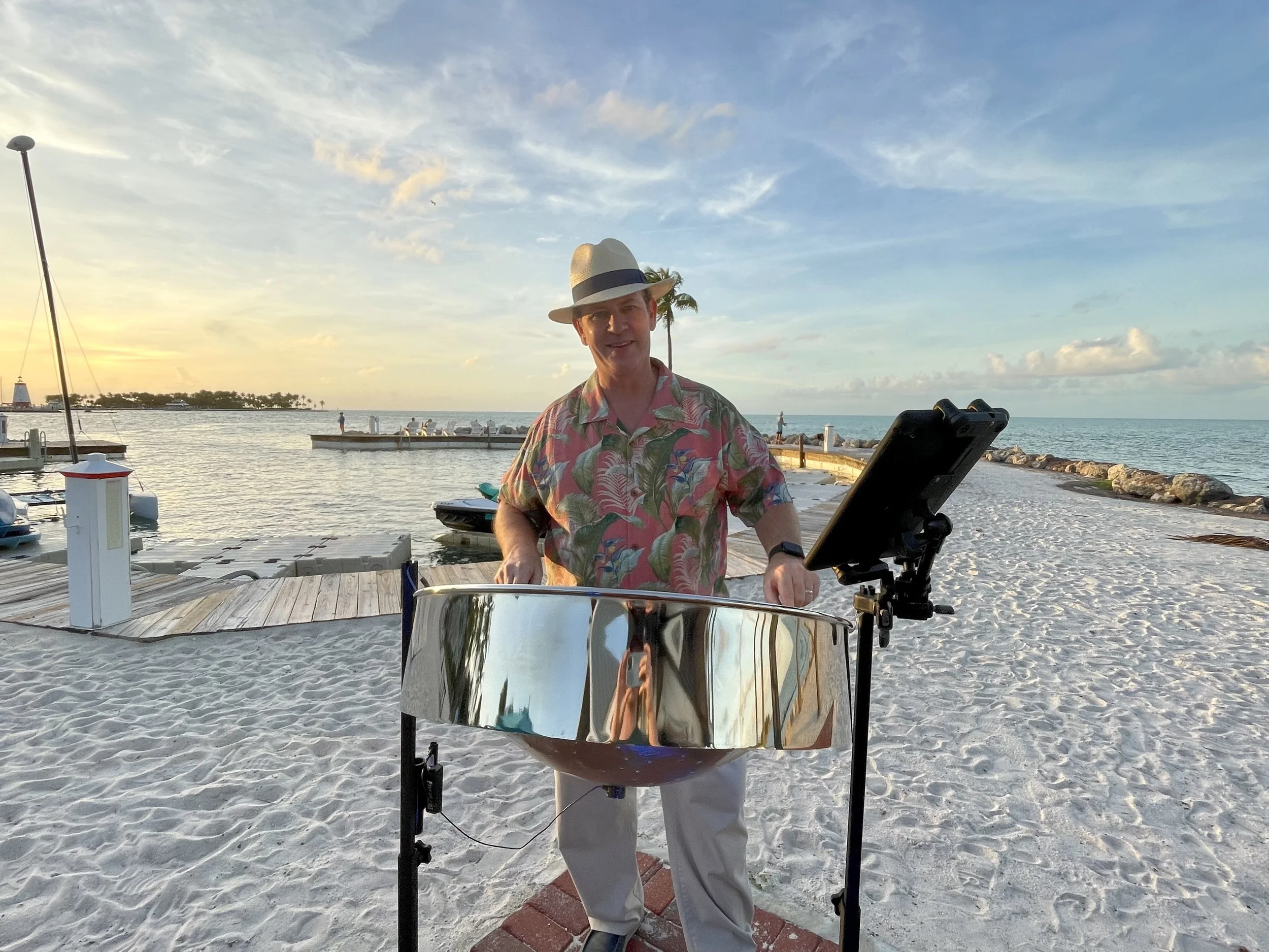 A man wearing a floral shirt and a fedora hat playing live steel drum music on a sandy beach during sunset, with a marina and calm ocean in the background. Tranquility Bay Beach Resort, Marathon Florida, Florida Keys.