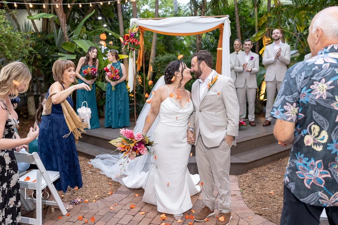 A newlywed couple shares a kiss under an outdoor wedding arch decorated with flowers, with friends and family celebrating around them in a lush garden setting. A Steel Drum Musician played. Hemingway House and Museum, Key West, Florida