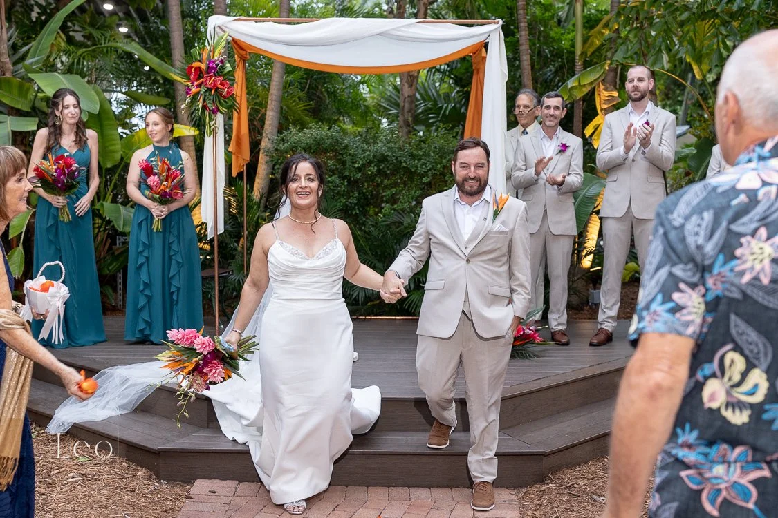 A couple in wedding attire holding hands and smiling during a wedding ceremony outdoors, with bridesmaids and groomsmen clapping behind them, and friends or family in the foreground. Hemingway House and Museum, Key West Florida