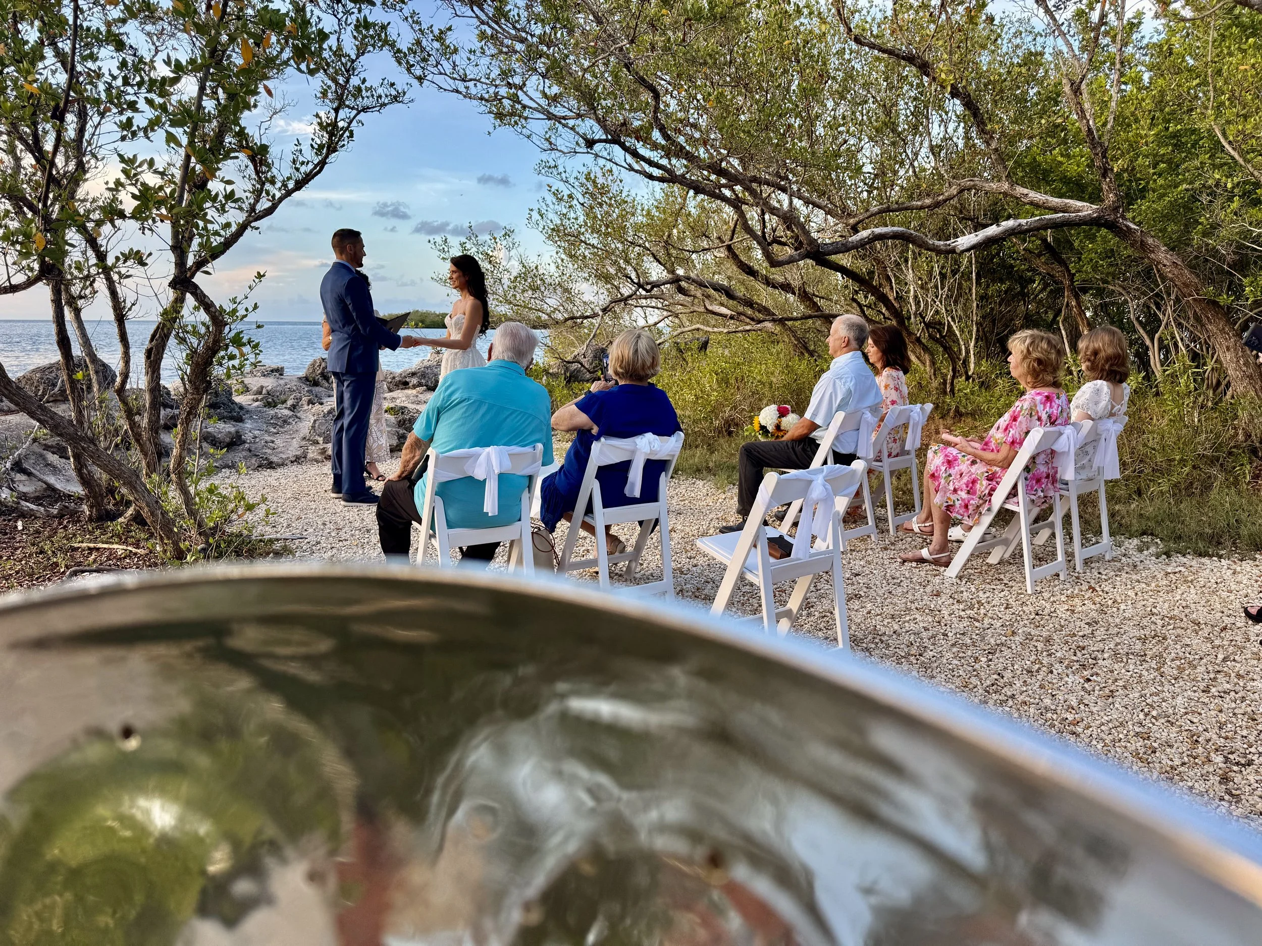 A beachside wedding ceremony with a couple exchanging vows, while live steel drum music is playing, guests seated on white chairs under trees, and the ocean in the background. Crane Point Hammock, Marathon Florida, Florida Keys, 