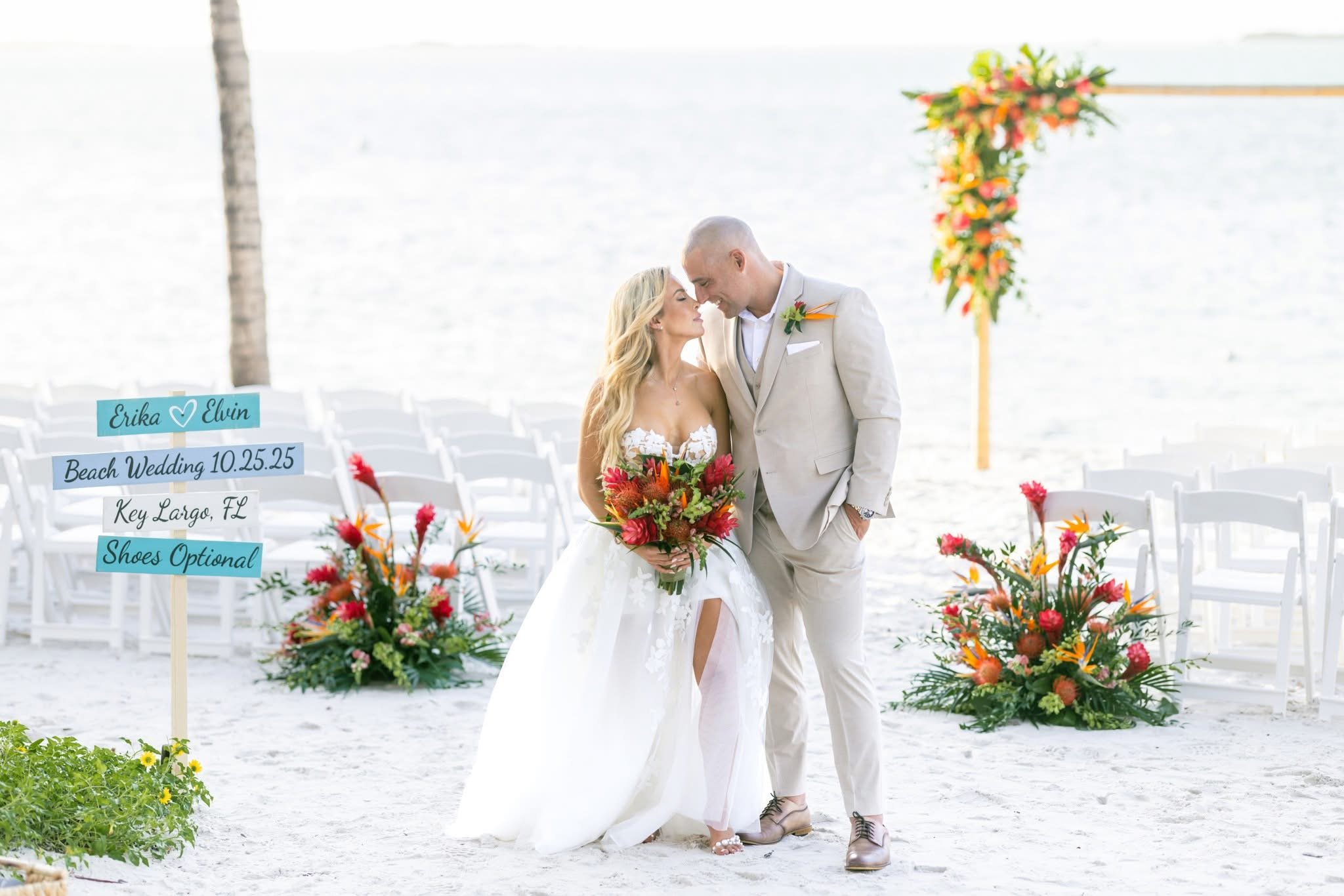 A bride and groom standing close together on a beach during their wedding ceremony, with a colorful floral arrangement and chairs in the background. Playalargo Beach Resort and Spa, Key Largo Florida, Florida Keys.