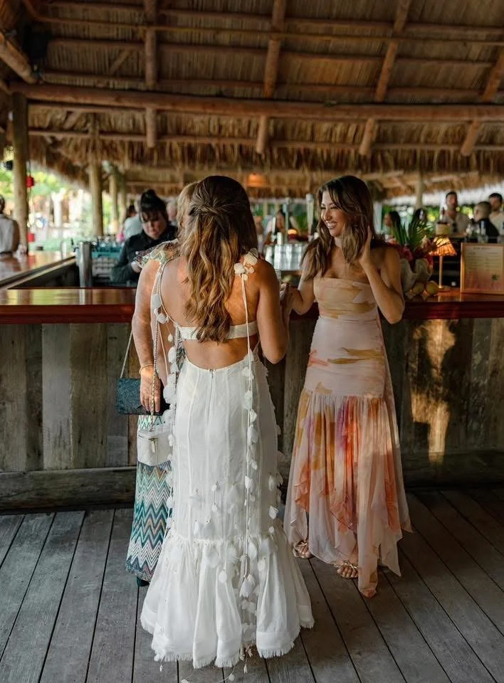 Three women in dresses standing and talking at a bar during a wedding welcome dinner, under a thatched roof in a tropical setting, with a bartender and other people in the background. Marker 88 Restaurant, Islamorada Florida, Florida Keys.
