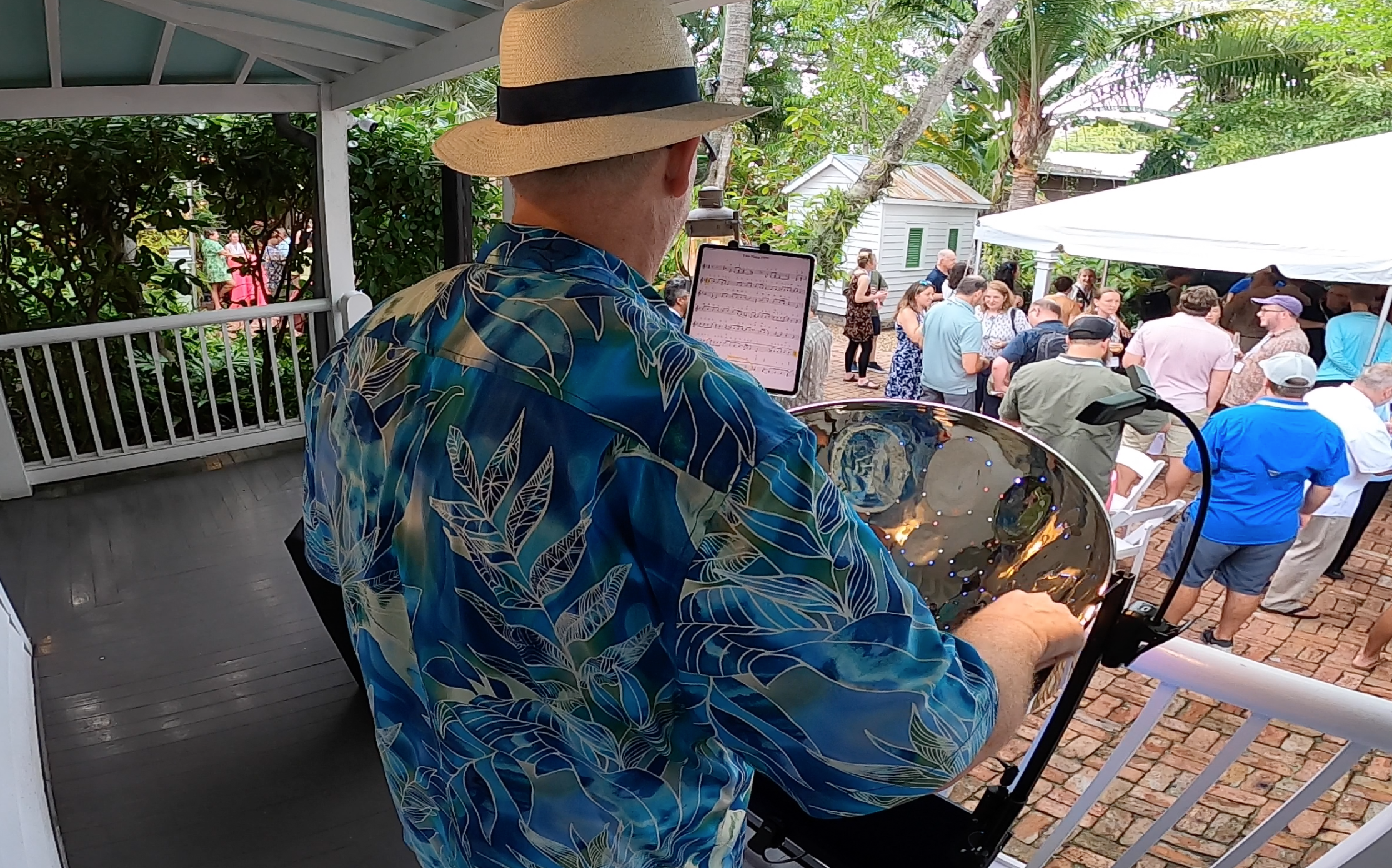 A man in a hawaiian shirt and straw hat playing a steel drum at an outdoor gathering for a corporate event with people socializing under tents and trees. Audubon House and Tropical Gardens, Key West Florida, Florida Keys Live Music