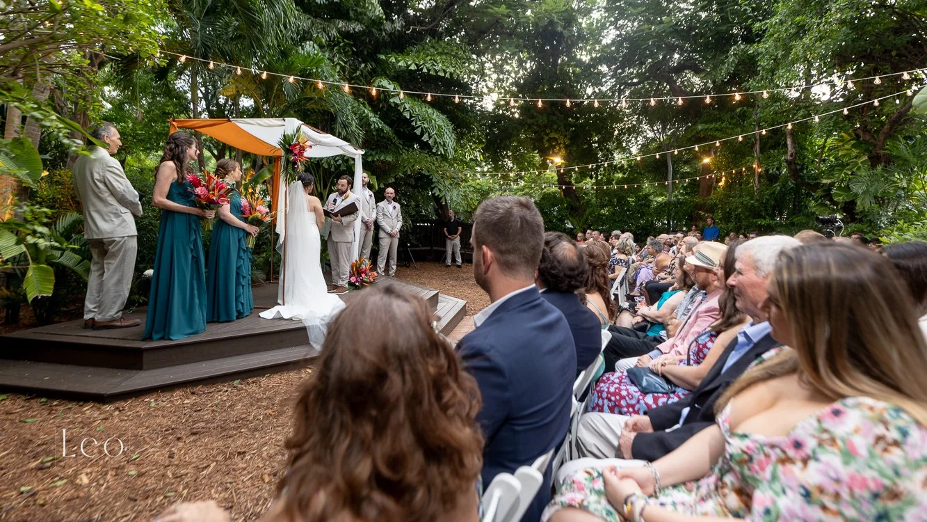 An outdoor wedding ceremony taking place in a lush, green garden with string lights overhead. The bride and groom stand on a small wooden platform, exchanging vows with bridesmaids and groomsmen nearby. Guests sit in white chairs, watching the ceremo