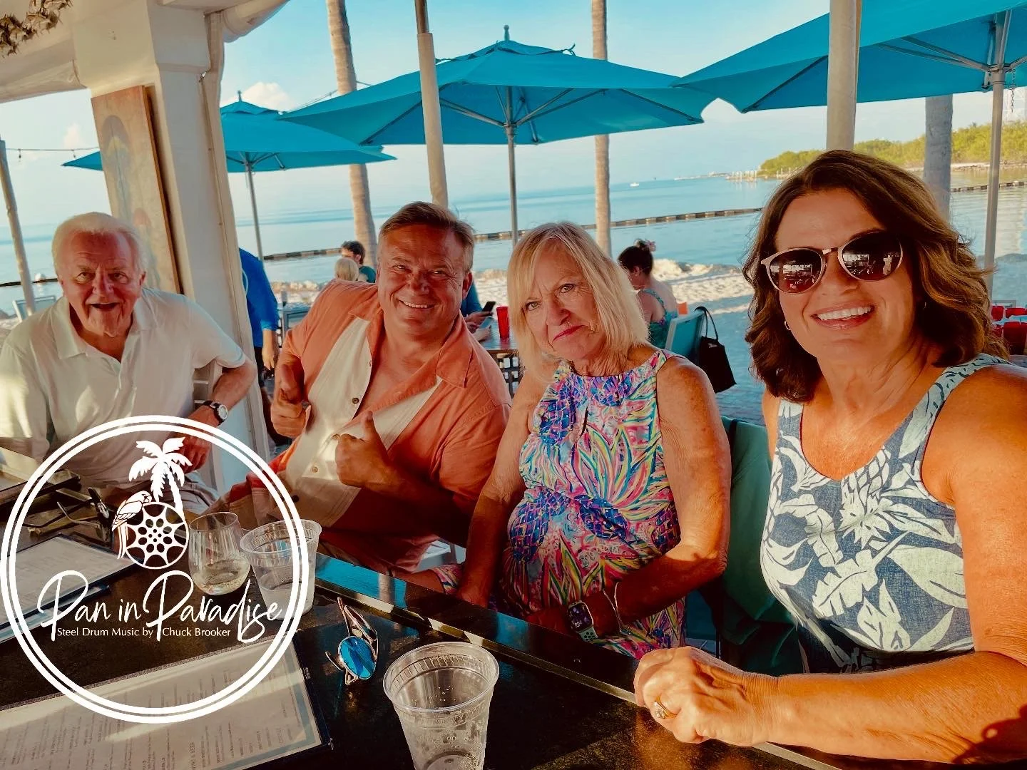 A group of four smiling adults sitting at a table outdoors near the beach, with blue umbrellas and palm trees in the background. Tranquility Bay Beachfront Resort, Marathon Florida.
