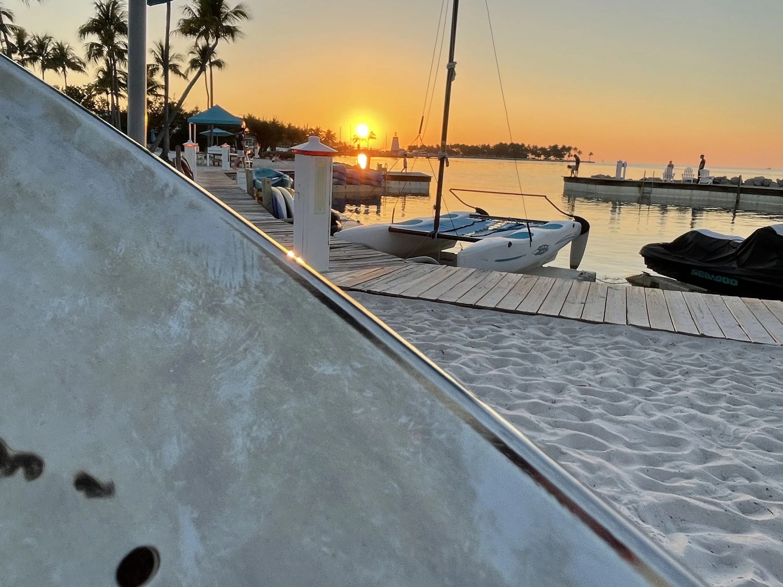 A sunset at a marina with boats docked, palm trees, and people walking along the pier, viewed from a sandy beach with a steel drum in the foreground. Tranquility Bay Beach Resort, Marathon Florida, Florida Keys