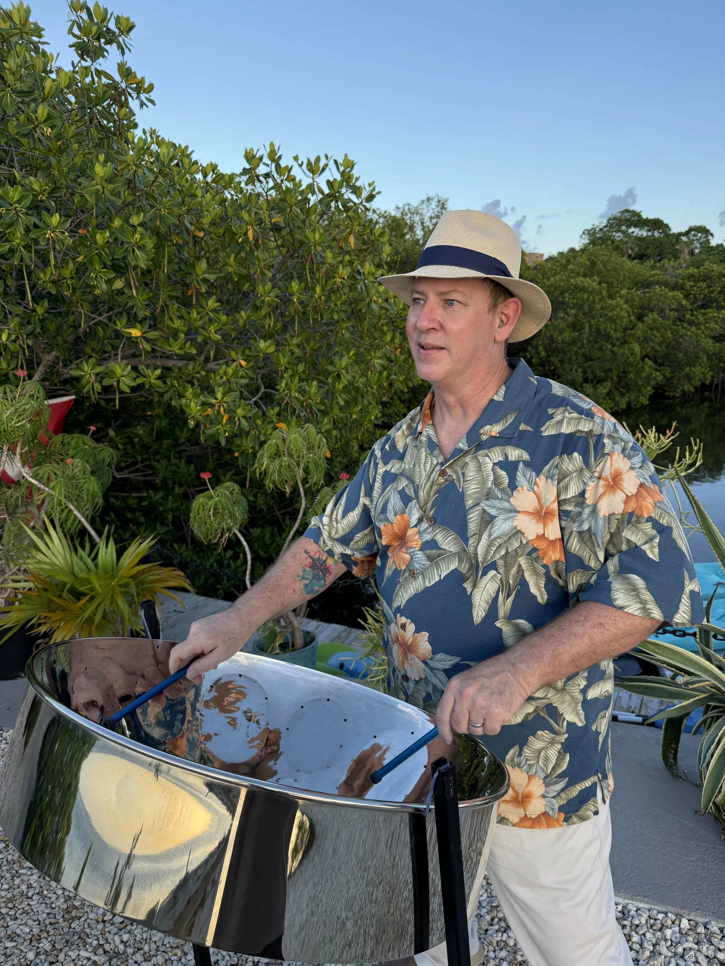 A man wearing a tropical shirt and a straw hat playing live steel drum music for a private event outdoors near water and green bushes. Crane Point Hammock, Marathon Florida, Florida Keys.