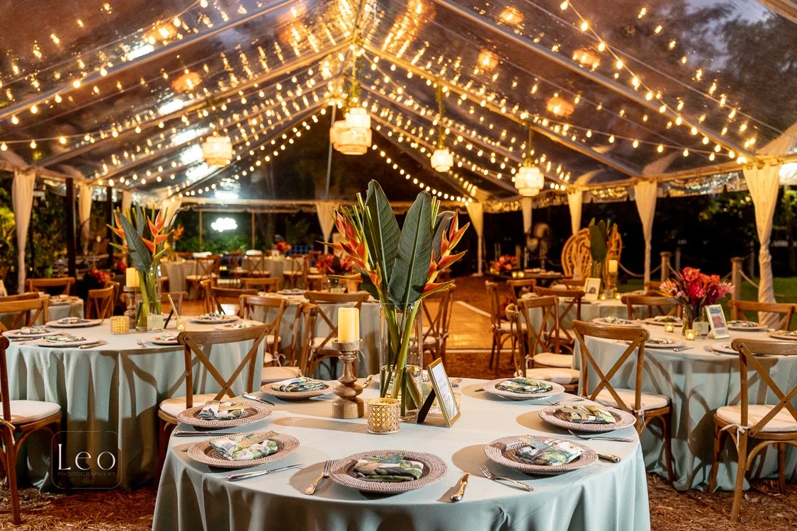Elegant outdoor event setup with round tables covered in white cloths, decorated with tropical flowers, candles, and plates, under a transparent tent adorned with string lights and lanterns. Hemingway House and Museum, Key West Florida
