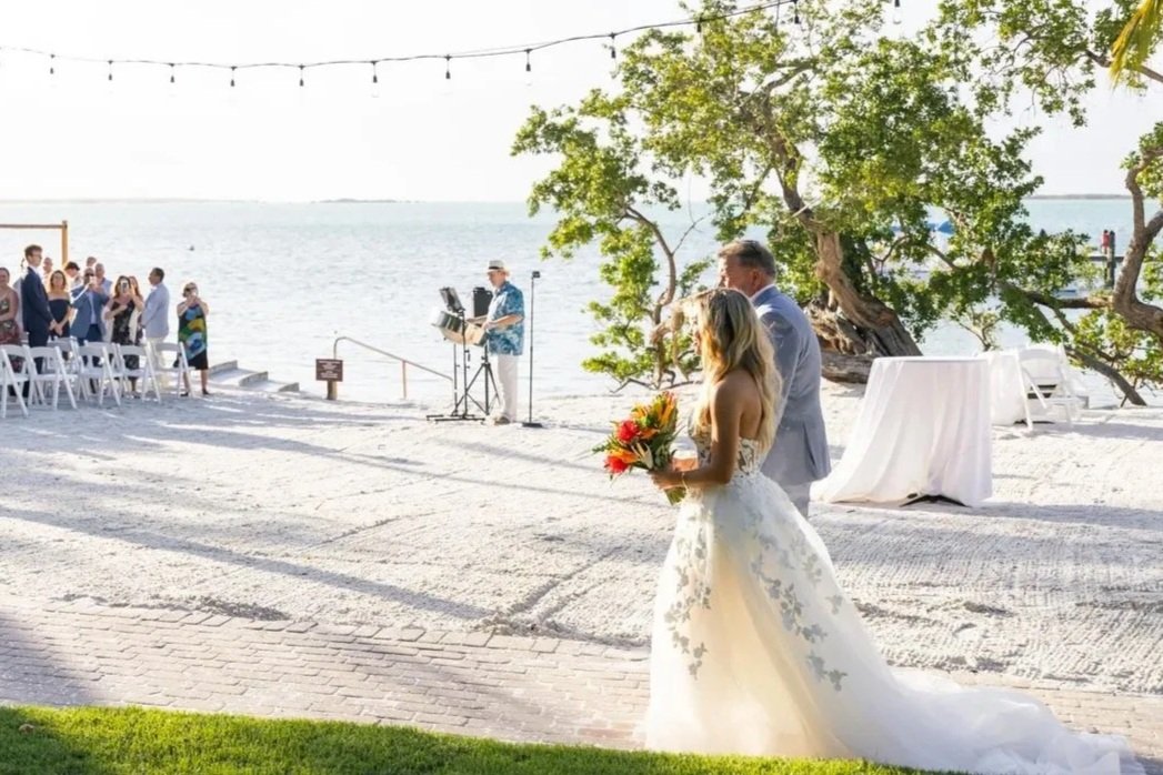 A bride in a white wedding dress holding a bouquet standing near a groom in a gray suit during an outdoor wedding ceremony by the water with a musician playing Steel Drum nearby. Playalargo Resort and Spa. Key Largo, Florida. In the Florida Keys