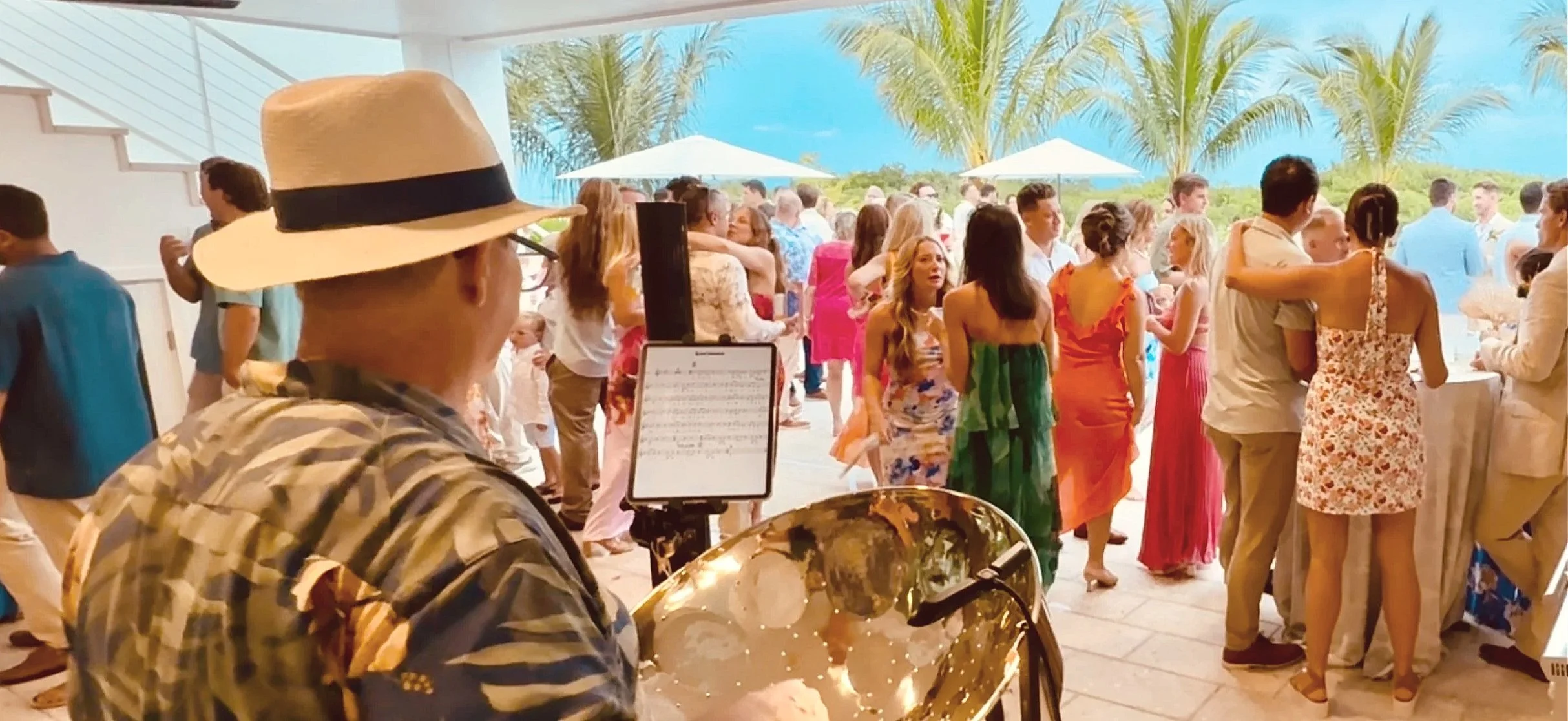 A musician wearing a large hat and sunglasses plays a steel drum at an outdoor wedding reception with guests in colorful dresses and tropical scenery in the background. Lunara Bay, Vacation Home Rentals, Key West Florida.