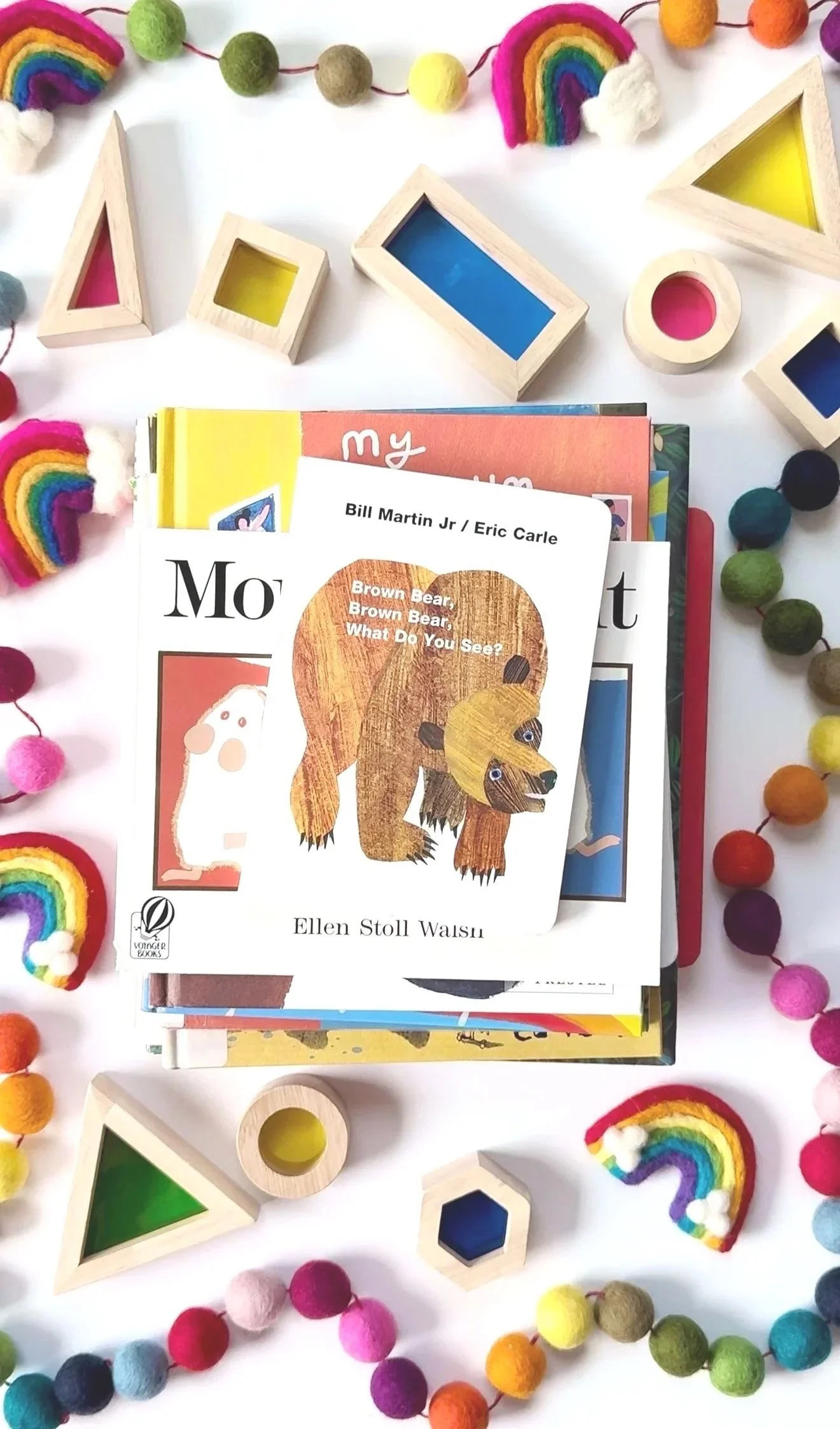 Children's books, including 'Brown Bear, Brown Bear, What Do You See?', surrounded by colorful felt rainbow and pom-pom decorations, with wooden geometric shape toys on a white surface.